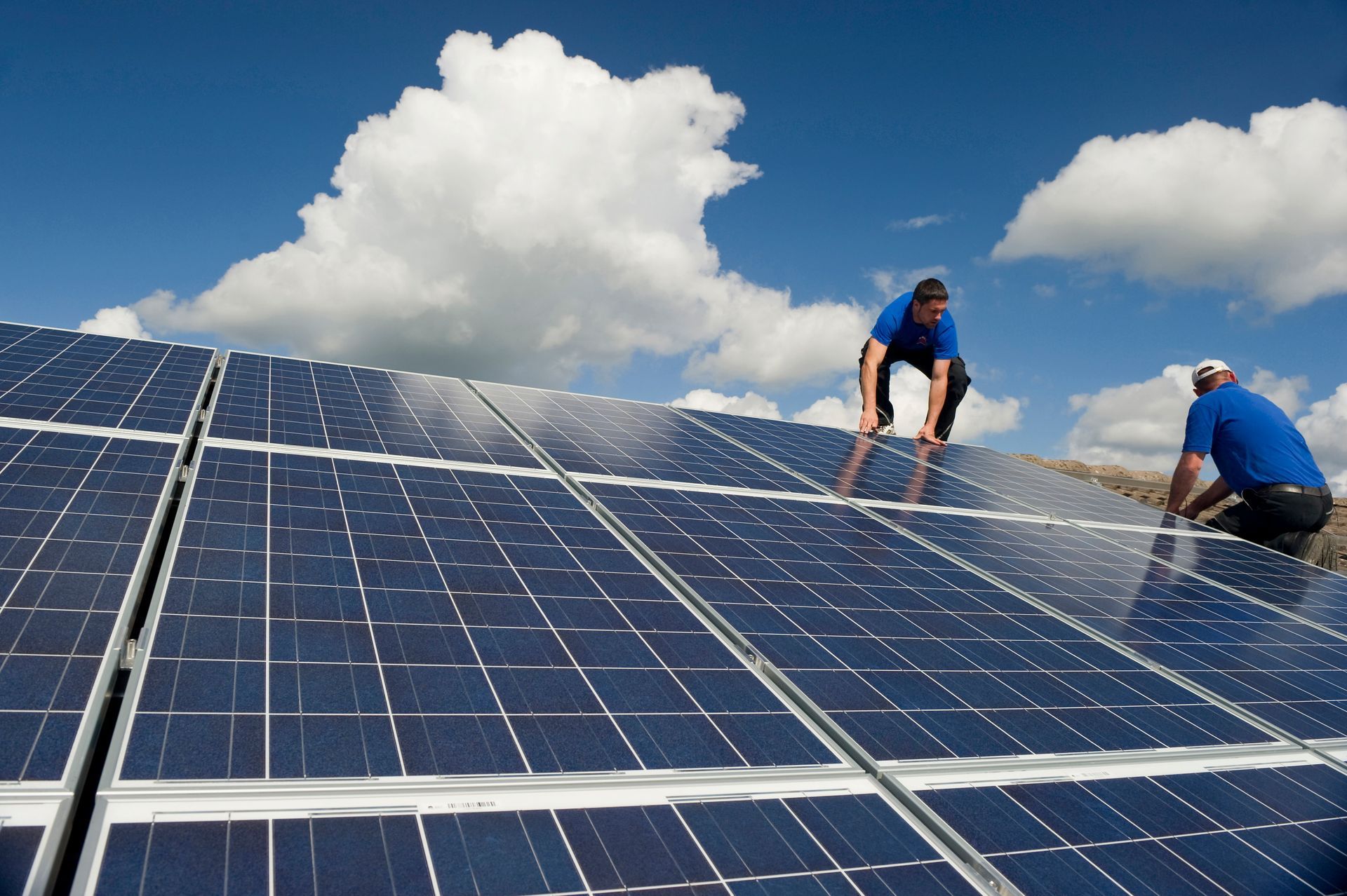 Workers installing solar panels on a roof against a blue sky with clouds.
