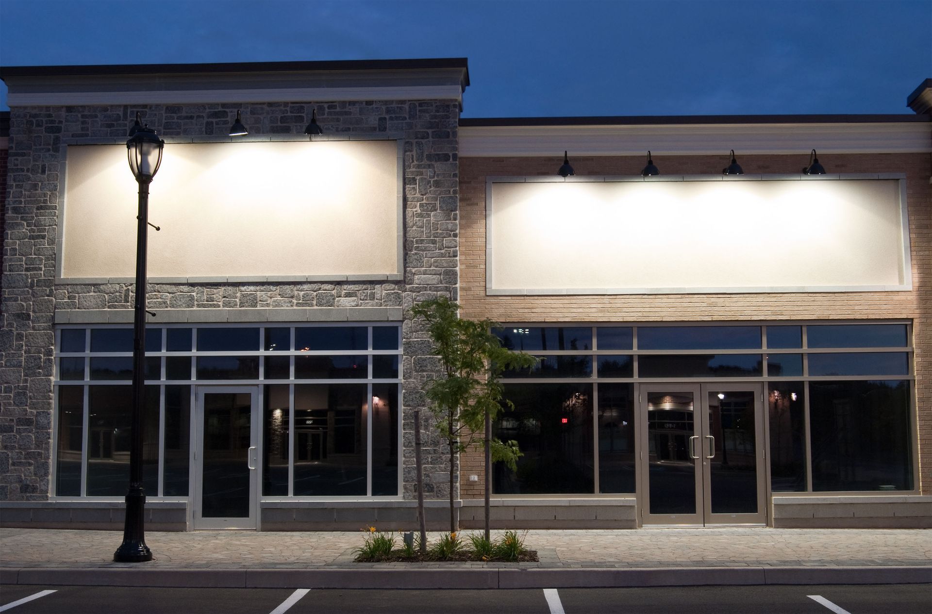 Storefronts at dusk, illuminated signage. Cobblestone street, lamp post, clear windows.