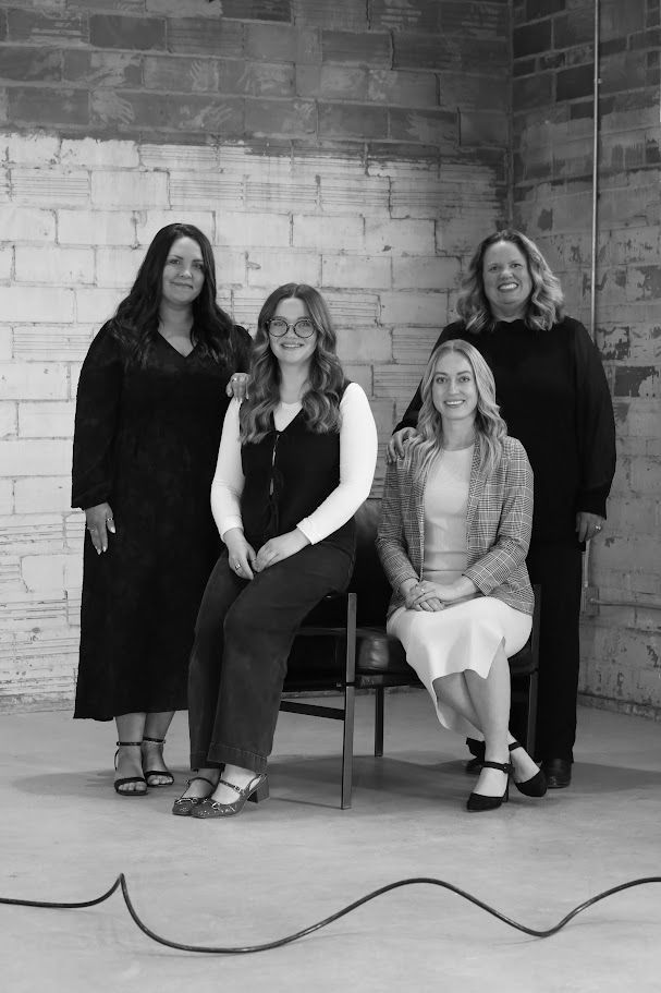 Three women posing in a rustic studio, two standing behind one seated in a chair.