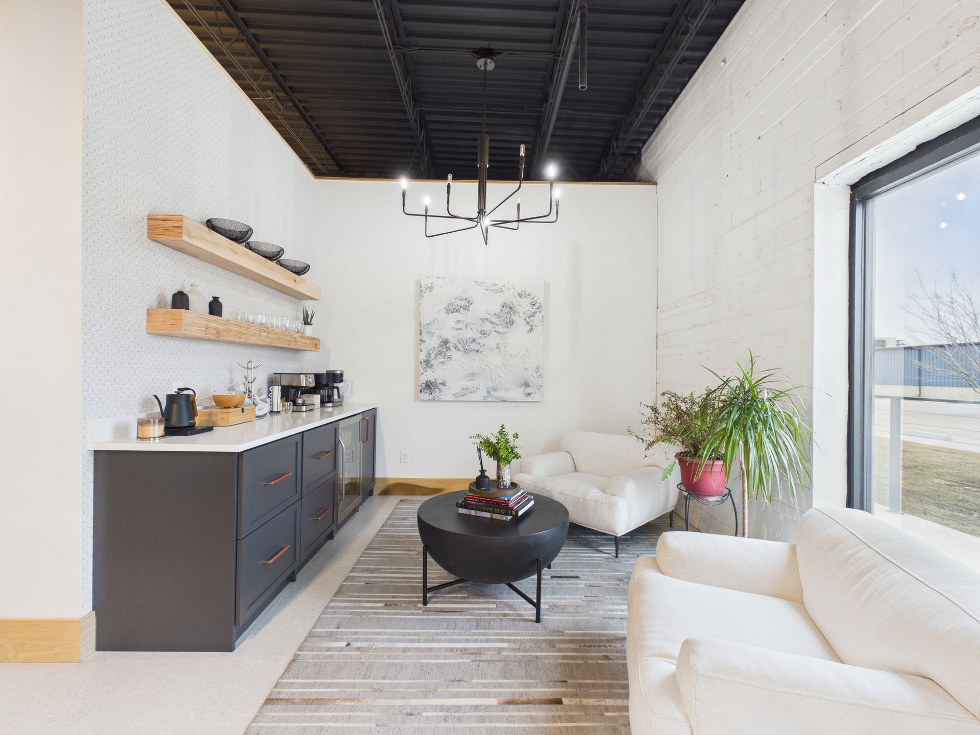 A modern lounge area with dark cabinets, floating shelves, a chandelier, seating, and a large window.