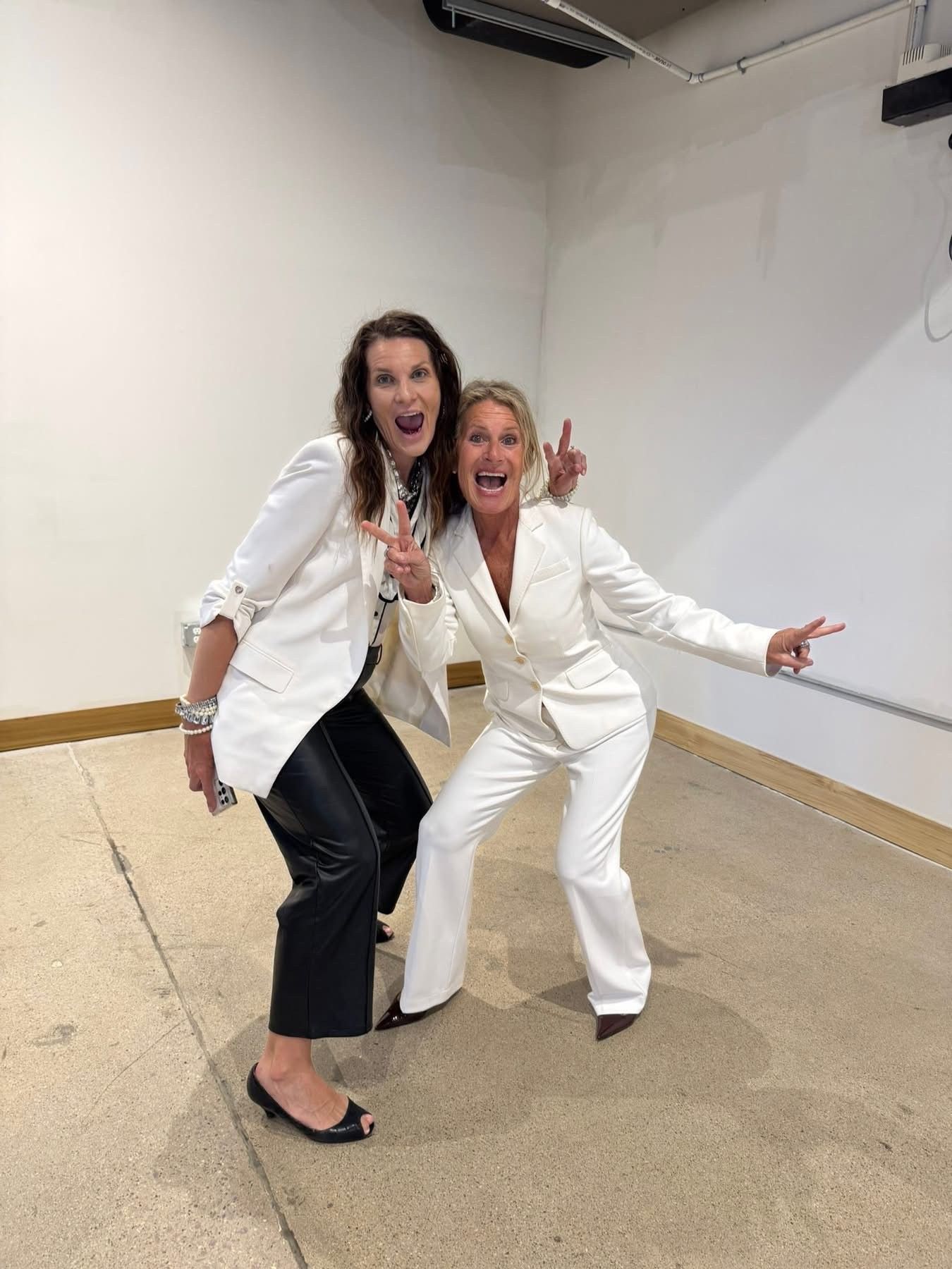 Two people in white suits pose playfully in a plain room, smiling and leaning toward the camera.