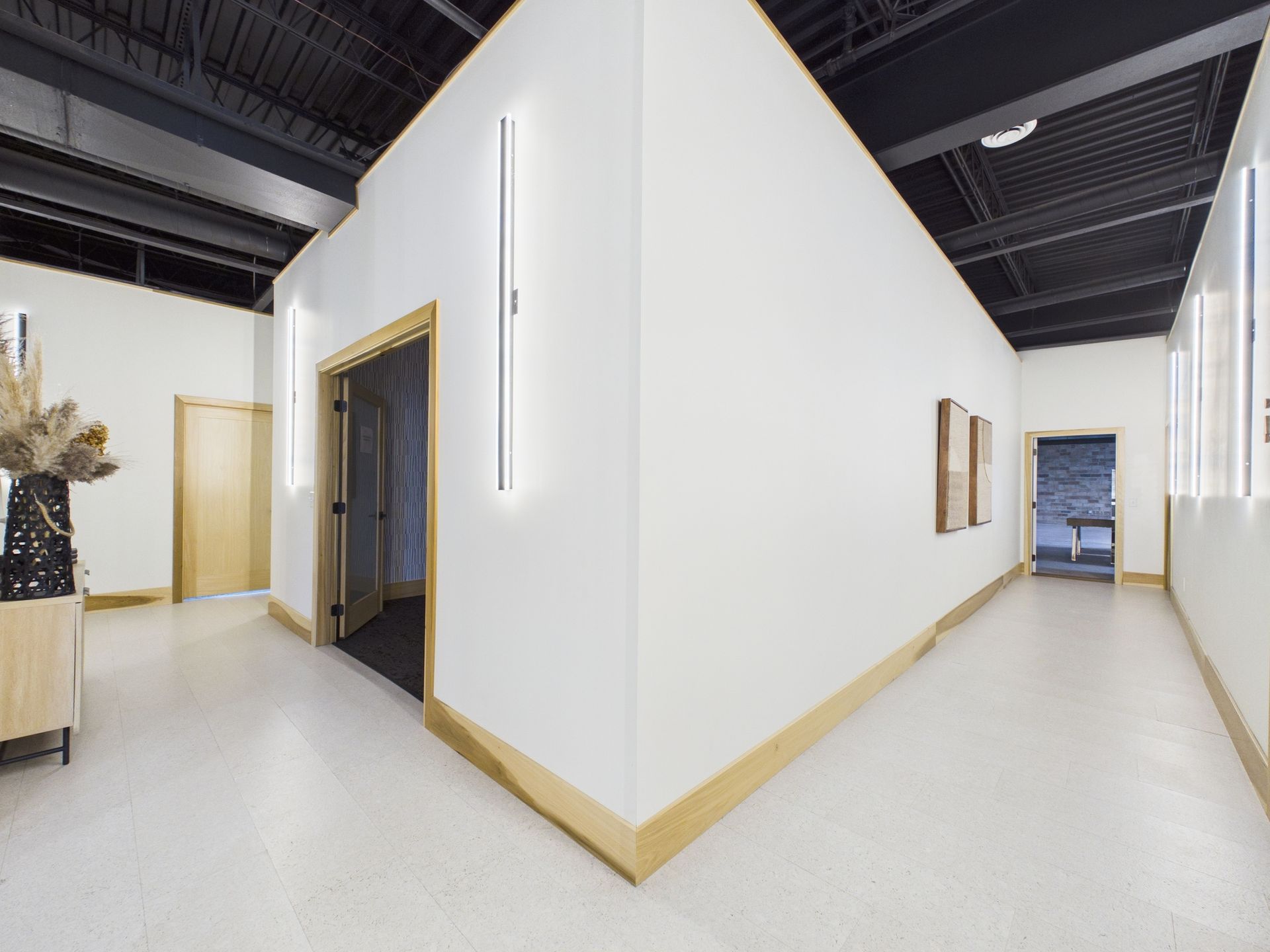 A minimalist hallway with white walls, blonde wood trim, vertical wall lights, and an open doorway under a black ceiling.