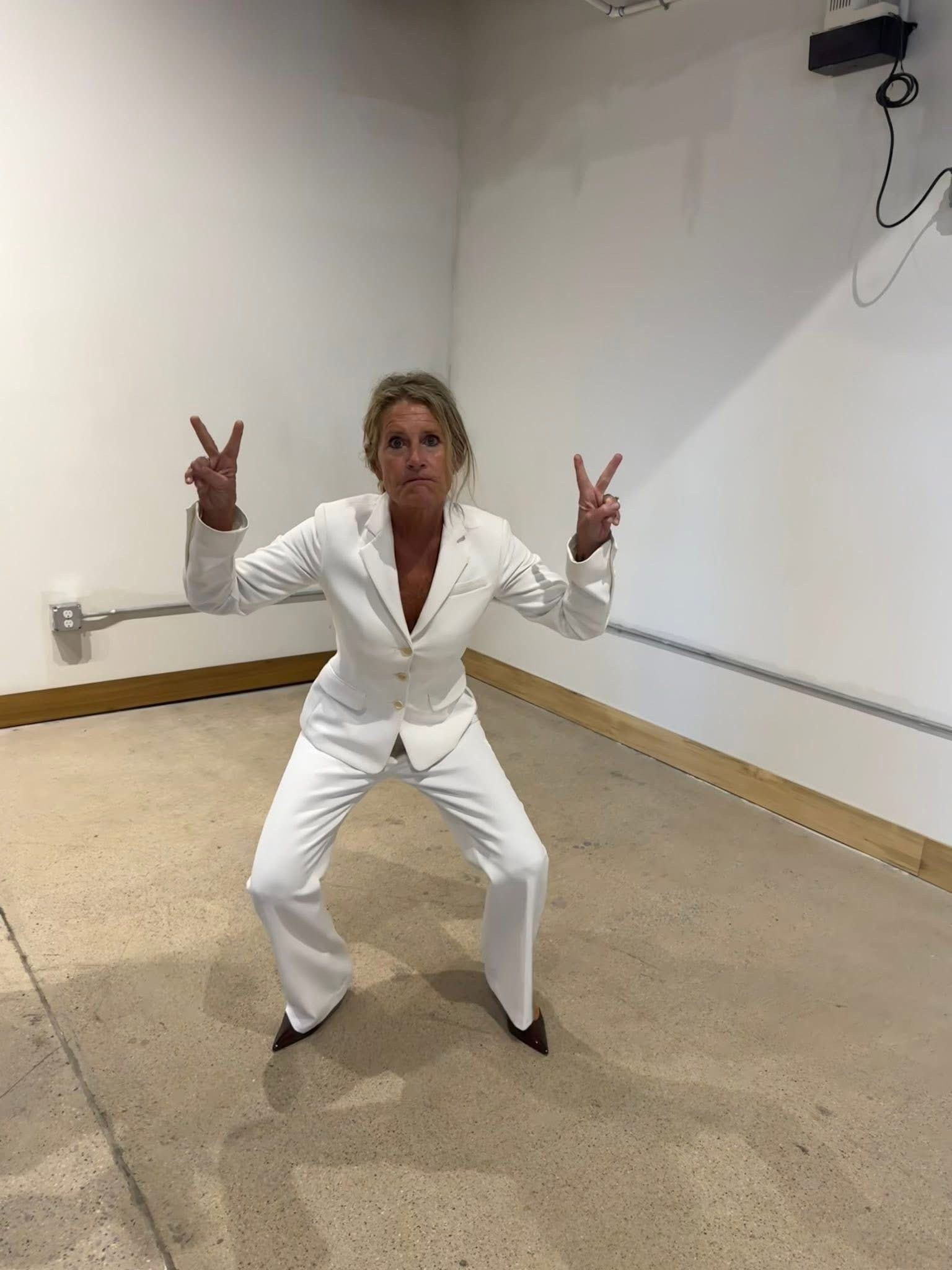 Person in a white suit crouches in a blank room, making peace signs with both hands.