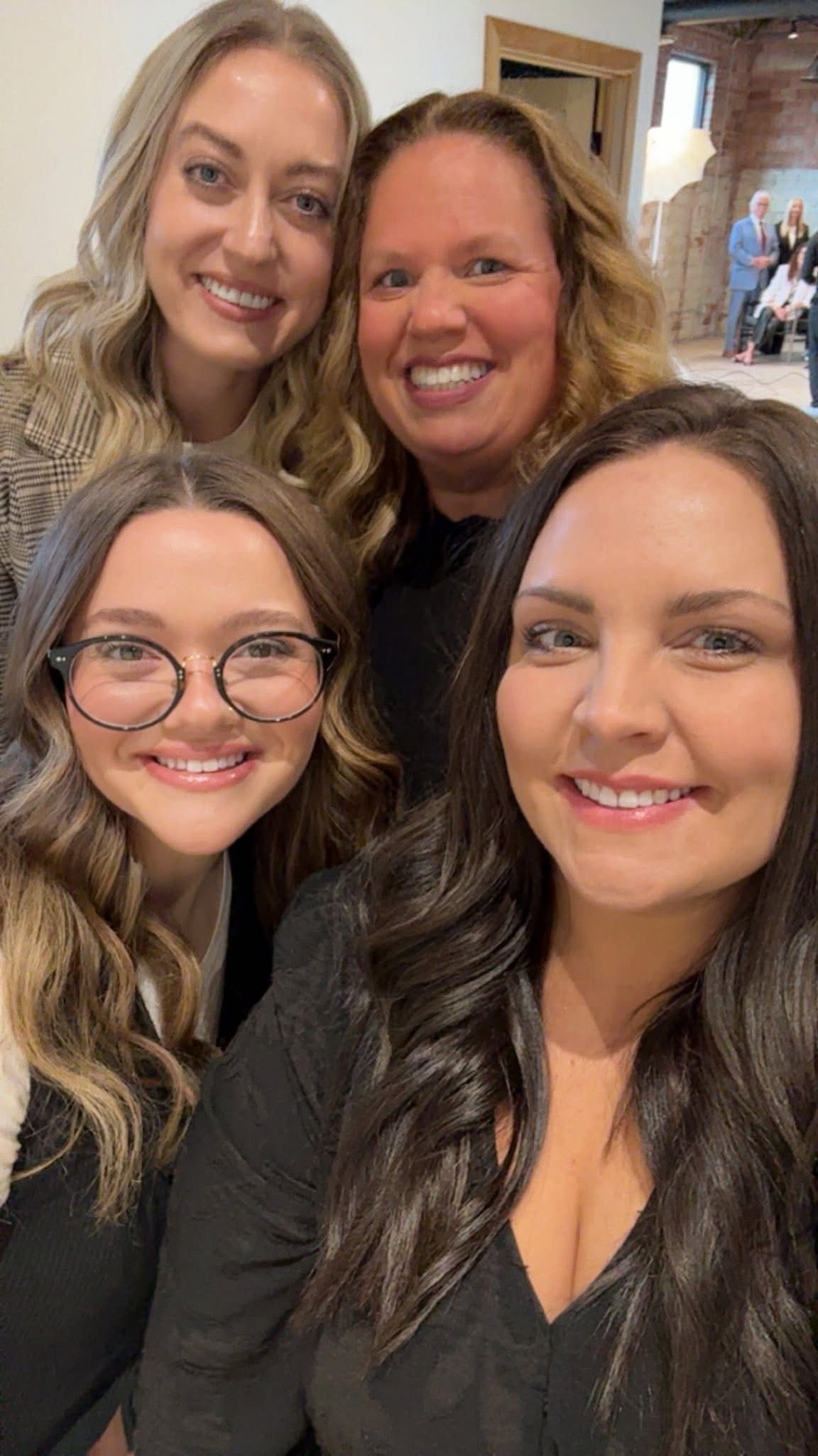 Four smiling people posing indoors for a selfie, with two in front and two behind.