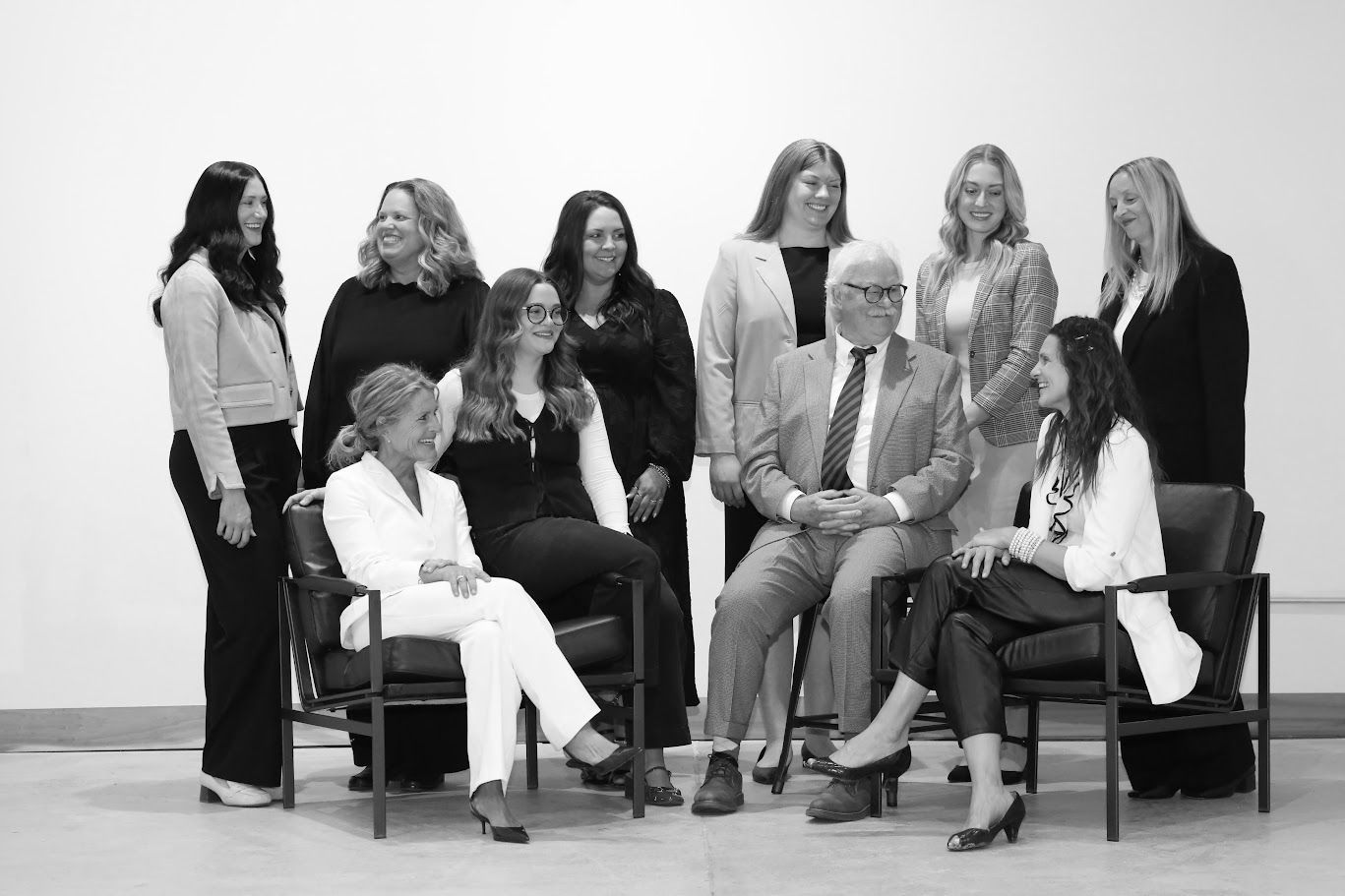 Group of people seated and standing in a studio portrait, smiling in light and dark outfits.