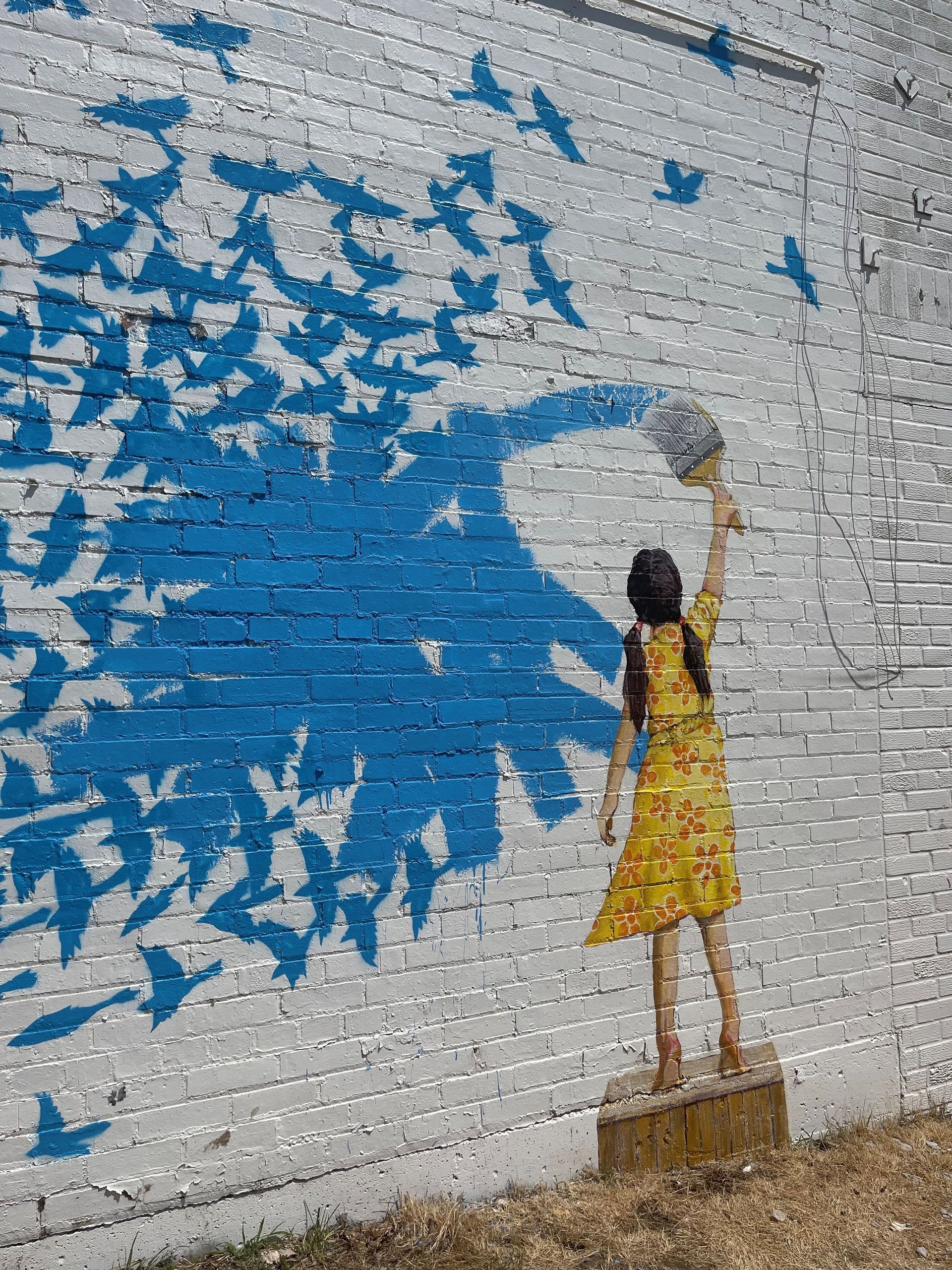 A mural of a girl releasing blue birds from a cage on a brick wall.