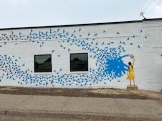 Mural of a person in yellow painting blue birds onto a white building's wall, birds swirling around the painter.