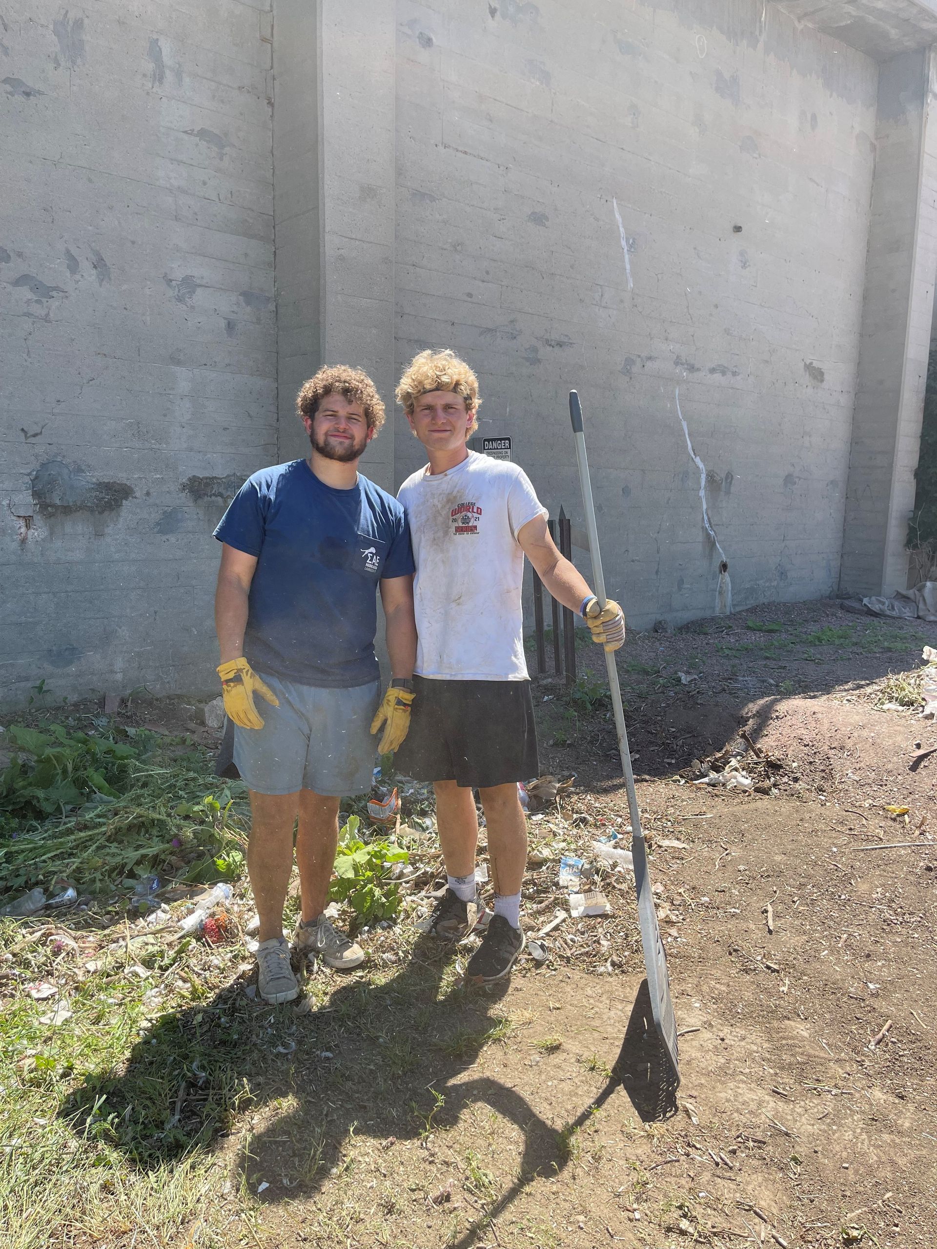 Two people stand by a concrete structure. One holds a long paddle. Both wear shorts, t-shirts, and work gloves.