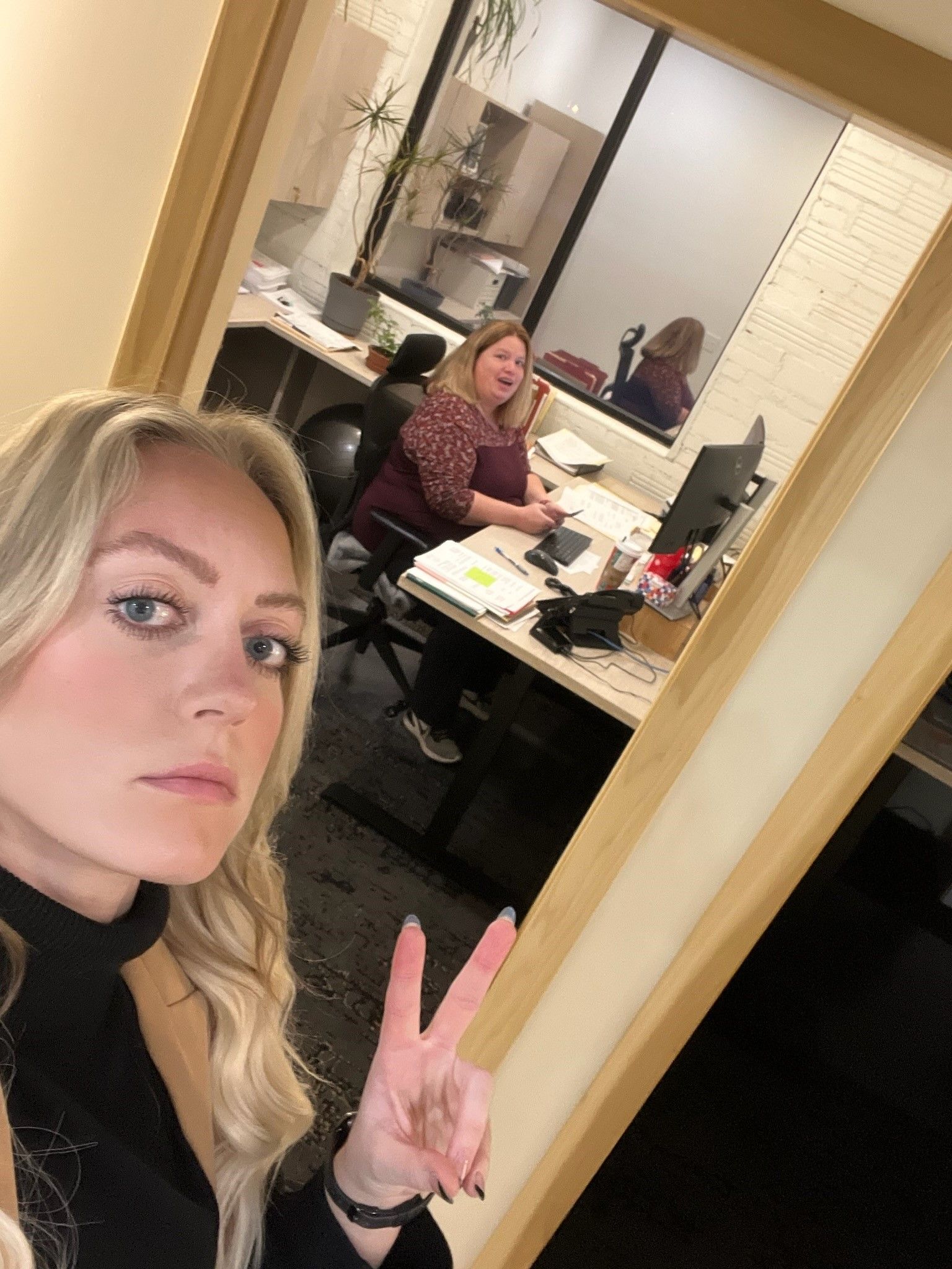 Blonde woman takes selfie, holding up peace sign. Another woman works at a desk through a glass office window.