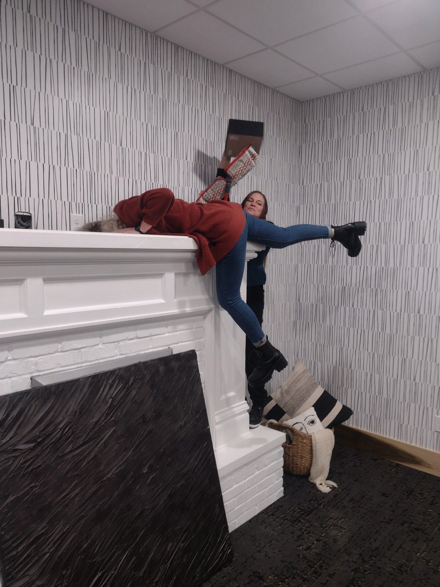 Two people playfully on a white fireplace. One reclined, other holding their leg up.  Black and white patterned wall.