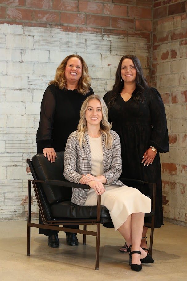 Three women pose in a studio, one seated in front of two standing behind her
