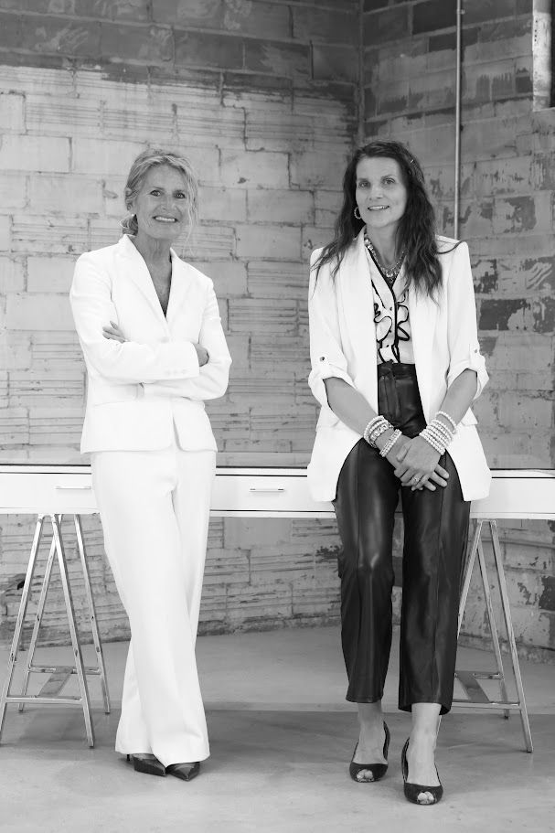 Two women standing in white outfits against a brick wall, arms crossed and hands clasped