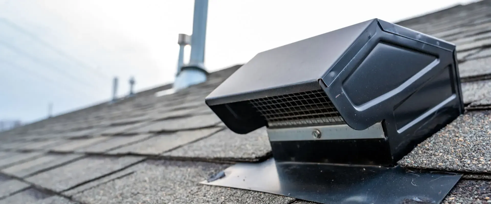 Black rooftop vent on asphalt shingles with distant rooftop background
