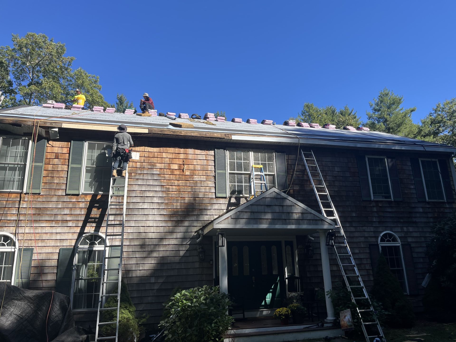 Workers on ladders repair a house roof under a clear blue sky.