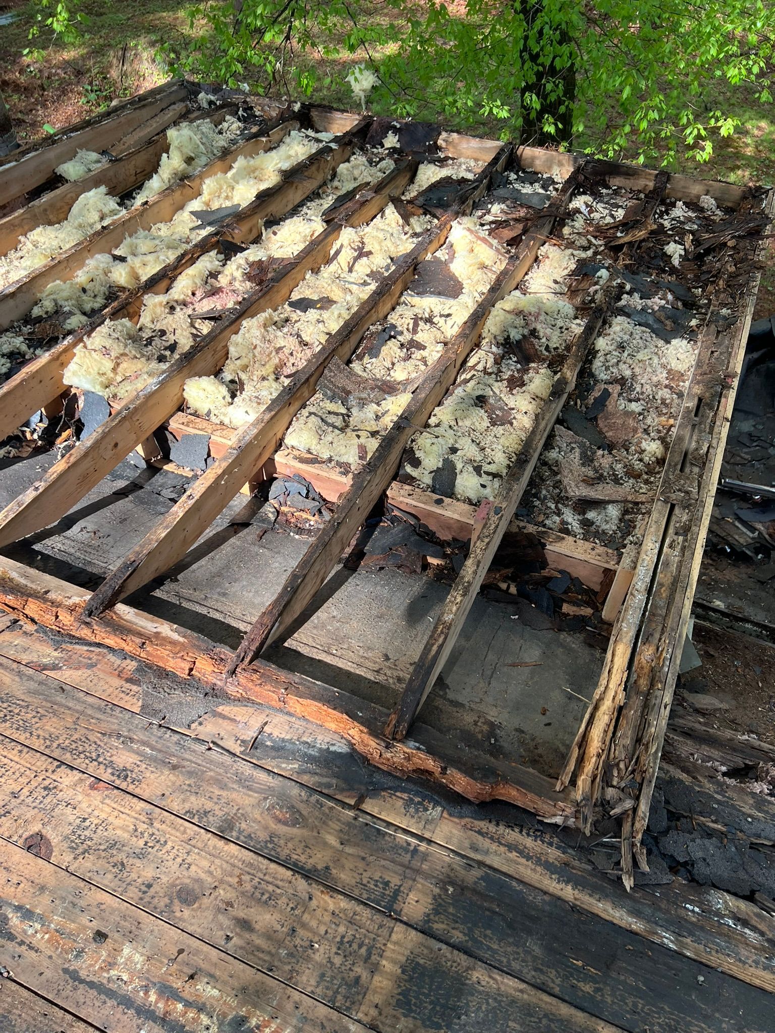Damaged roof with missing shingles and exposed beams, seen from above near trees.