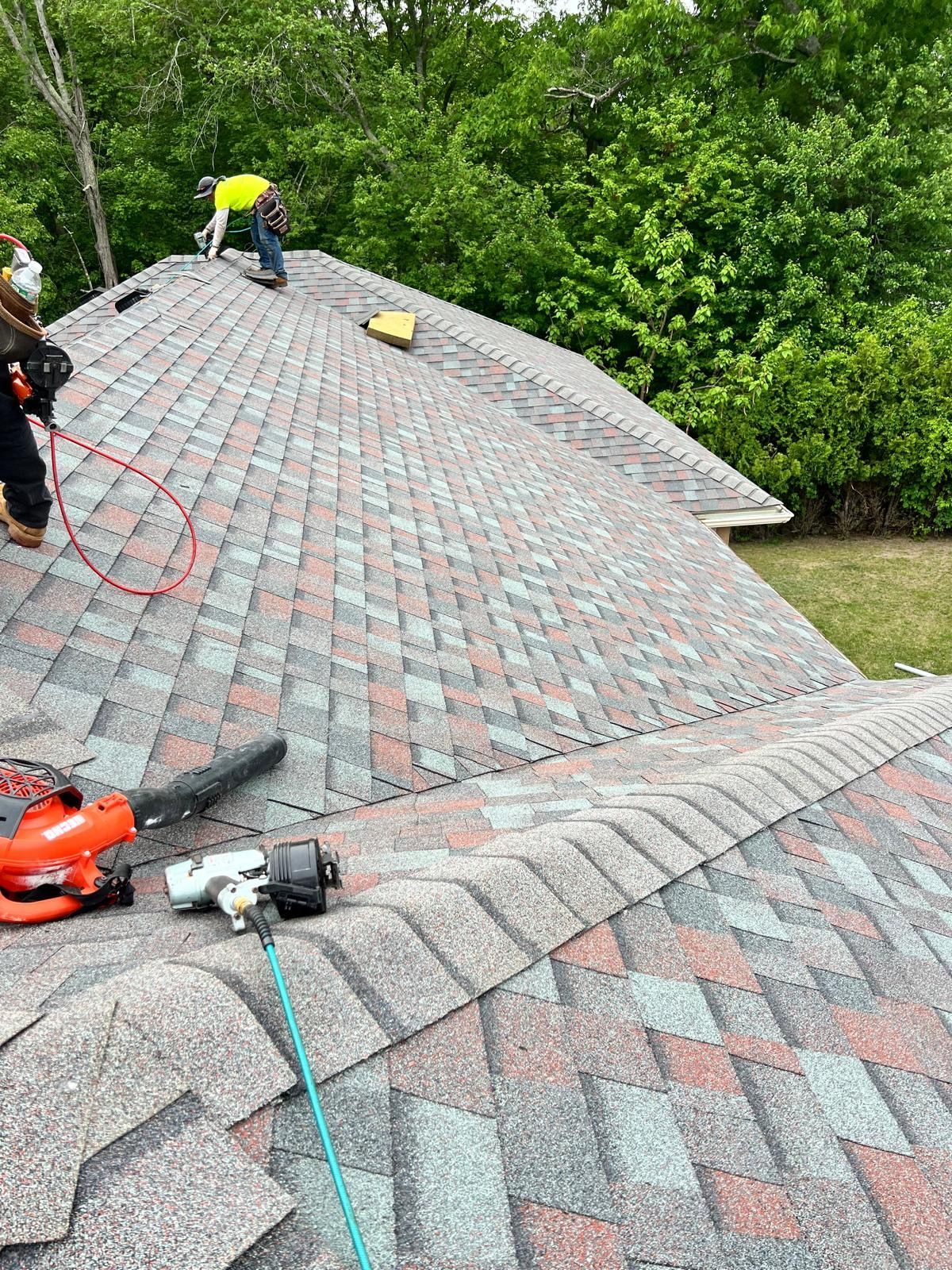 Workers repairing a gray shingled roof beside green trees, with tools and safety ropes visible.