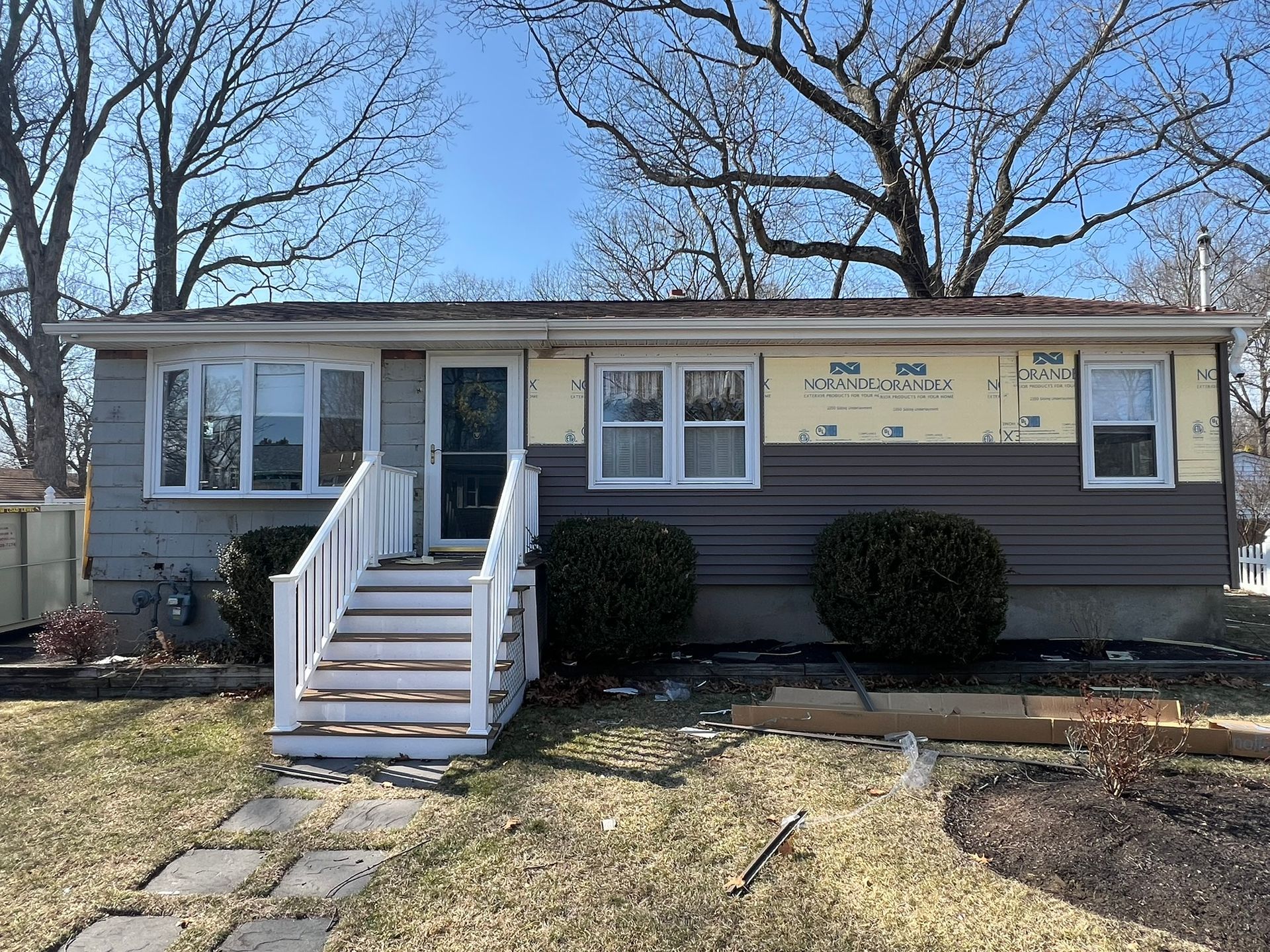 Single-story house with white steps, gray siding, and unfinished yellow exterior under clear blue sky