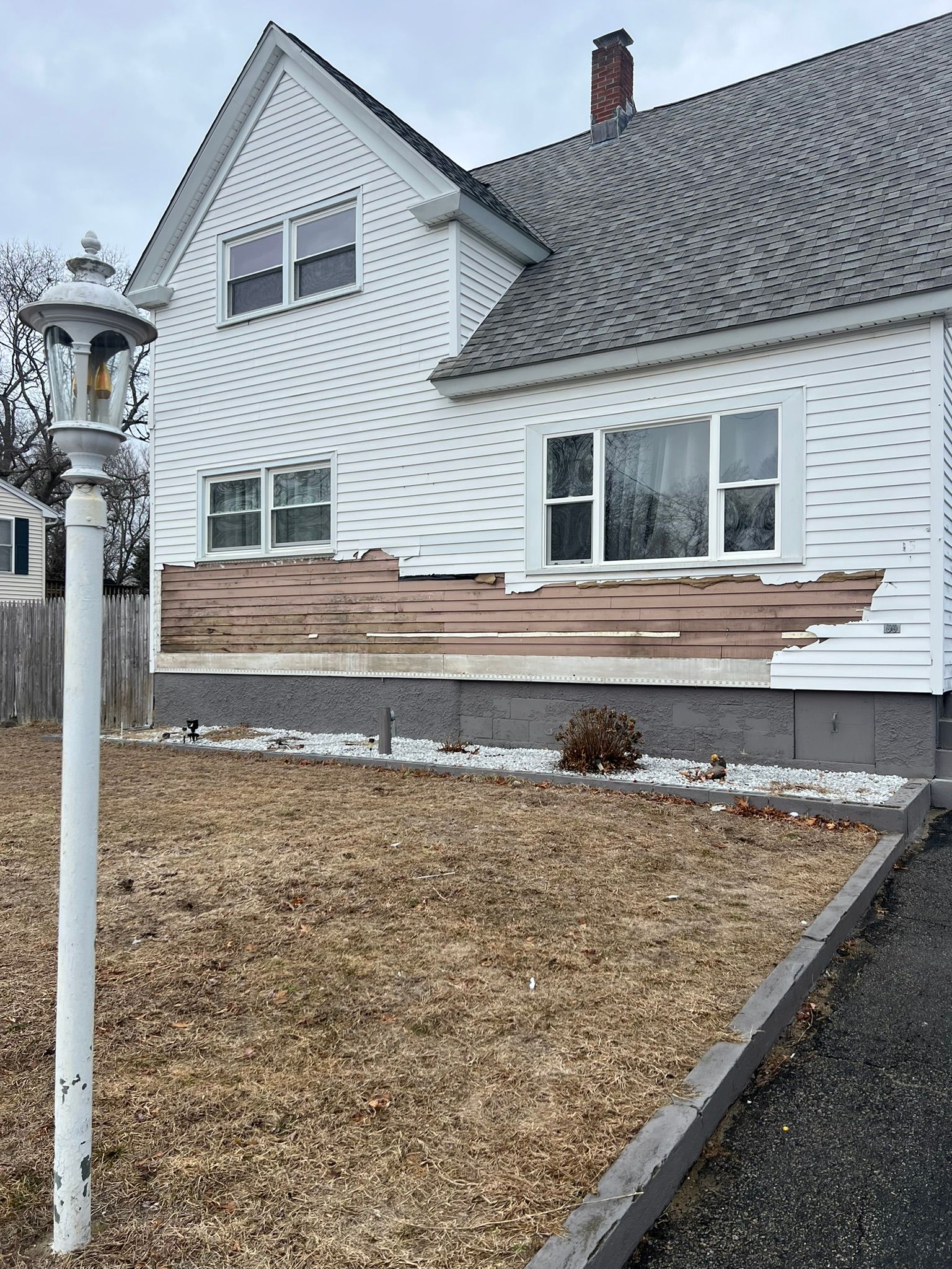 White house with damaged siding and a small front yard in winter; white lamp post in foreground