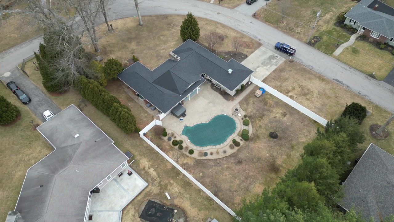 Aerial view of a house with a backyard pool, fenced yard, driveway, and neighboring homes.