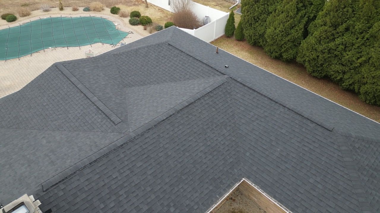 Aerial view of a gray shingled roof beside a backyard pool and tall hedges