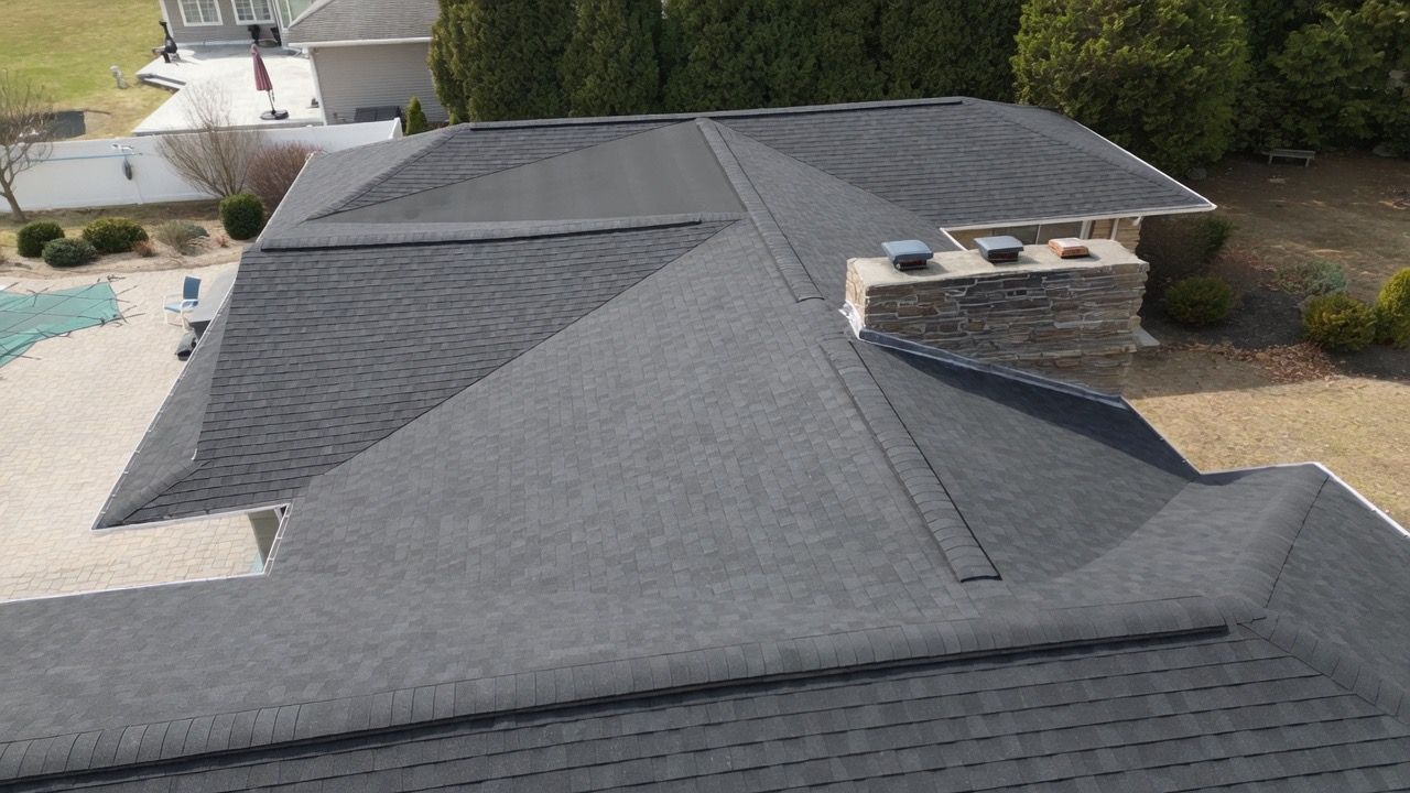 Aerial view of a dark gray shingle roof with a small chimney and neighboring houses in the background