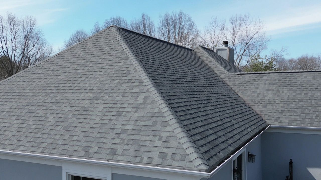 Gray shingled roof with a steep hip roof and dormer under a clear sky, trees in the background