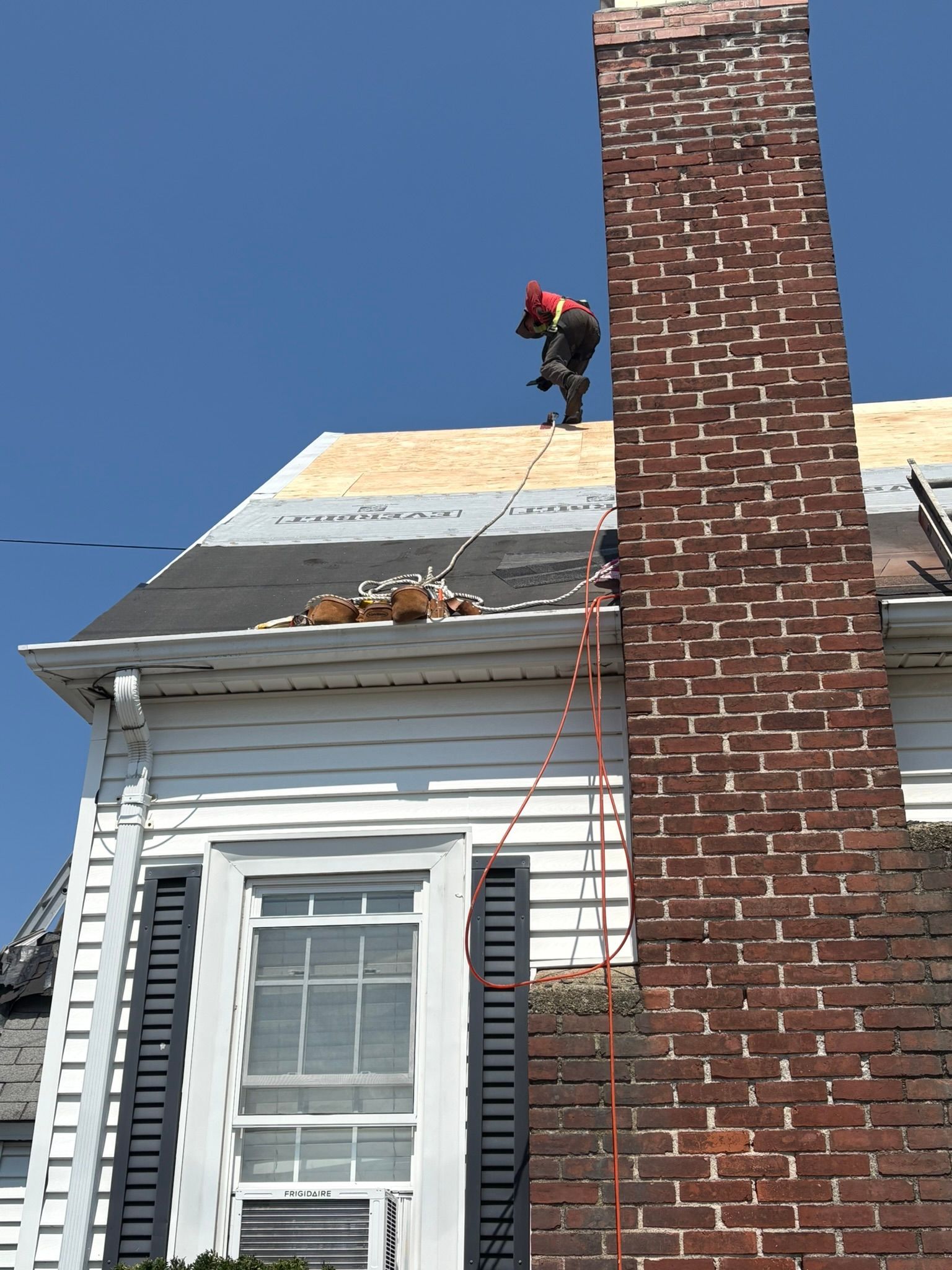 Worker on a house roof near a brick chimney, repairing shingles under a clear blue sky.