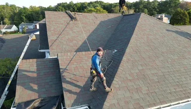 Worker on a steep shingled roof using a safety harness and hose.