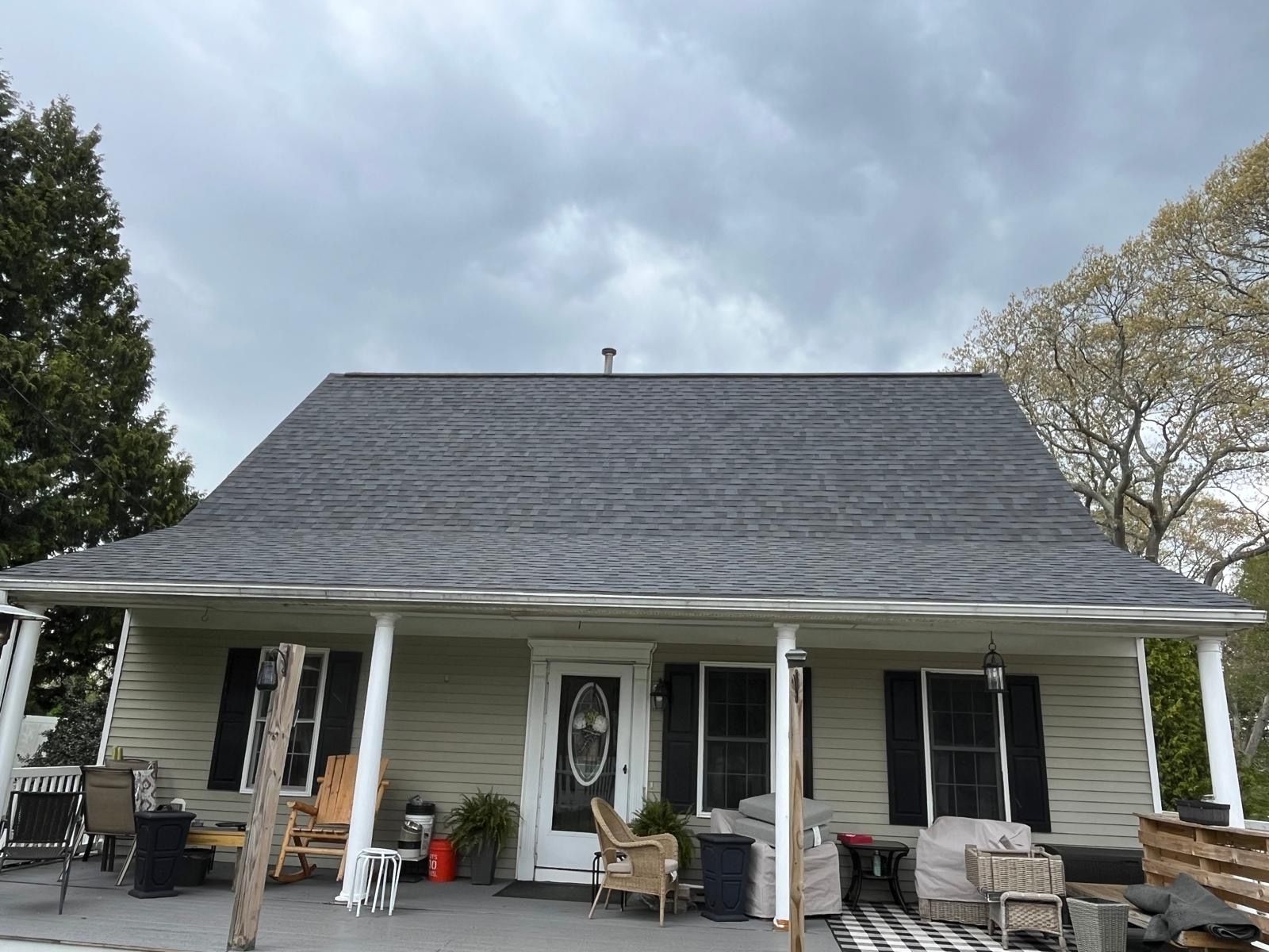 White house with a wide front porch, dark roof, and outdoor seating under a cloudy sky.