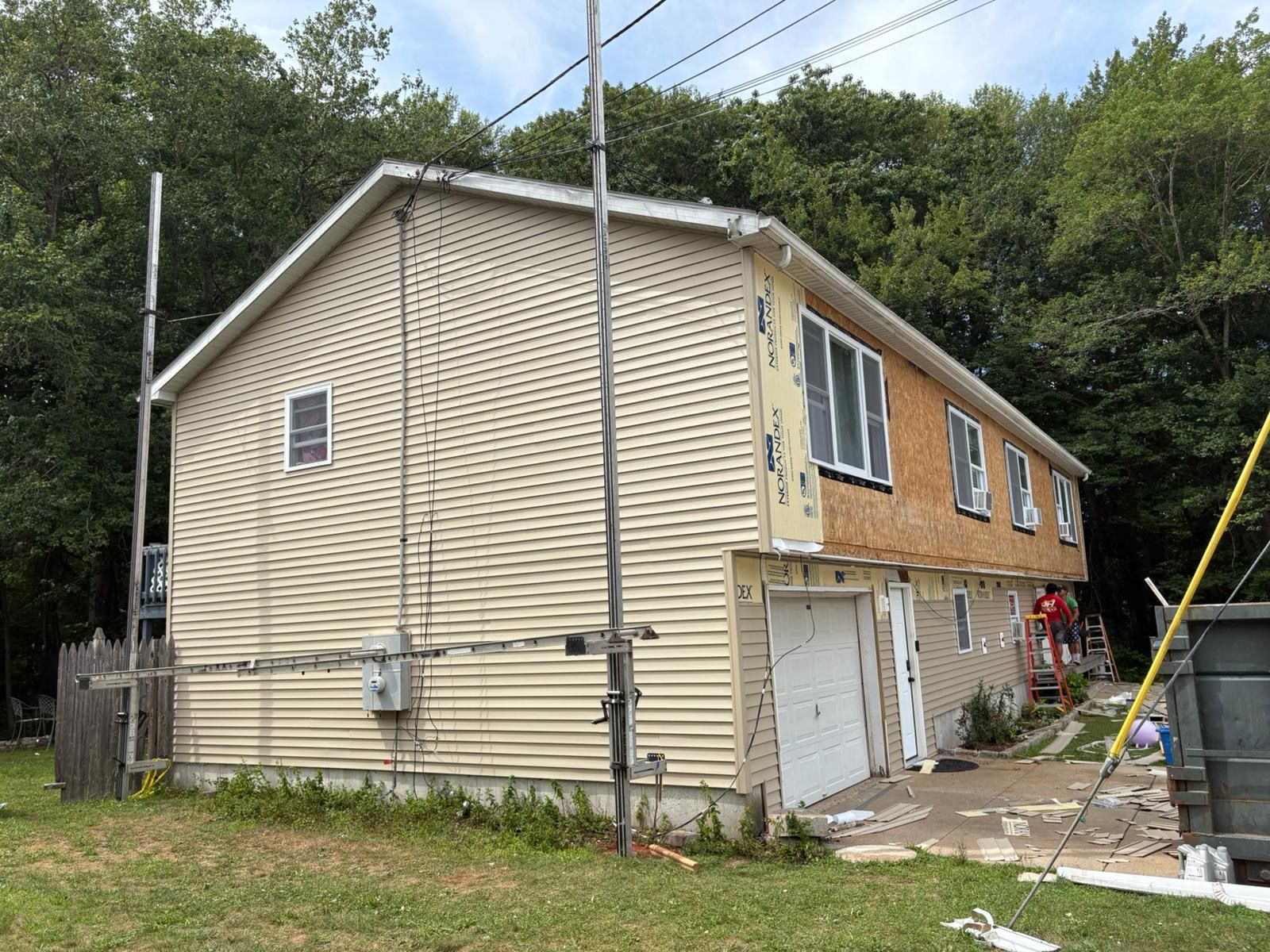 Two-story house with tan siding and white garage doors beside a wooded hillside