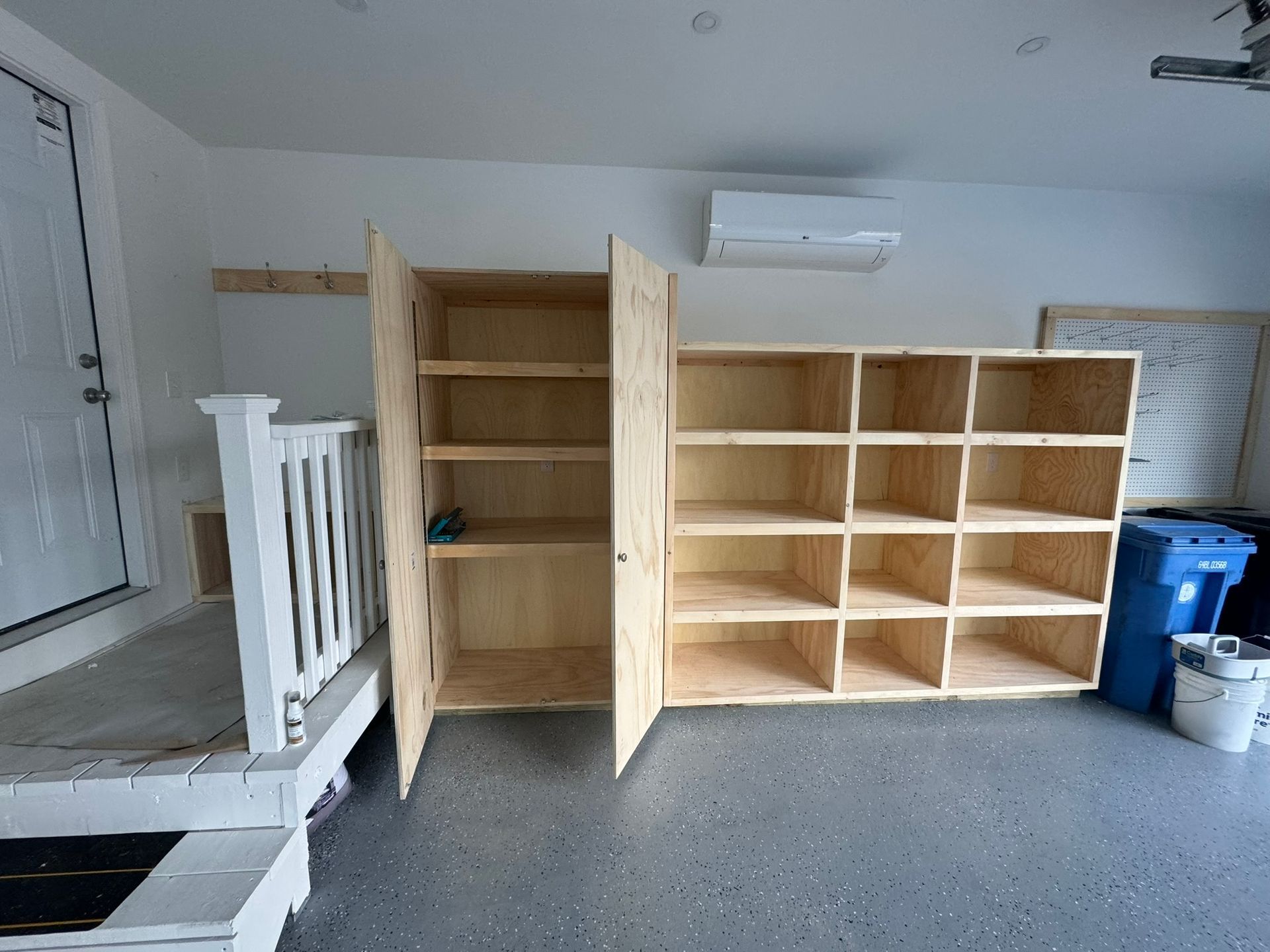 Built-in wooden shelving and cubbies in a garage, with a white railing and blue trash bins nearby