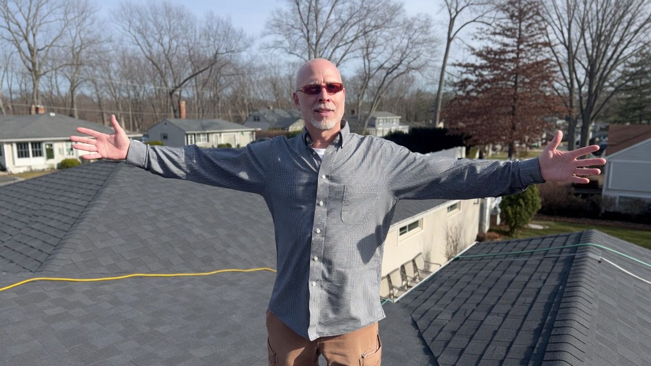 Person with arms outstretched on a rooftop, wearing sunglasses and a gray shirt, with trees and houses behind.