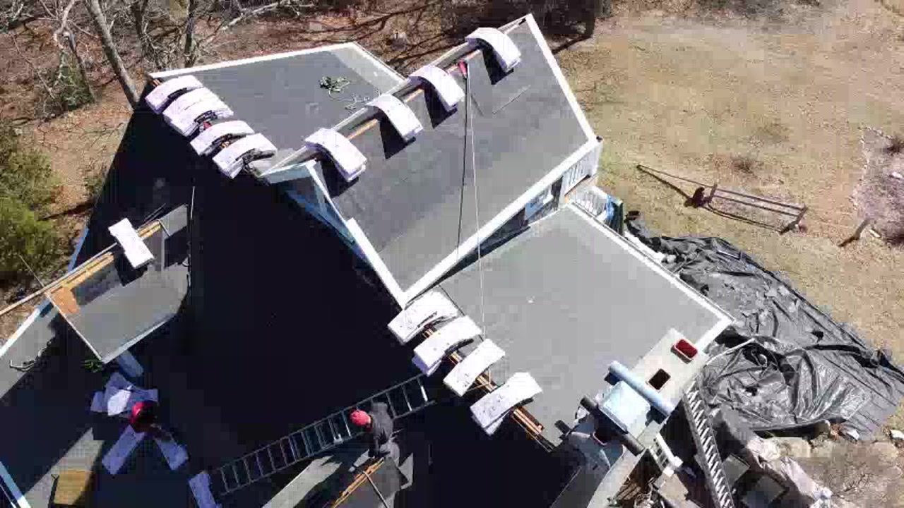 Aerial view of a house roof with dark gray shingles and several loose white panels or boards on top.