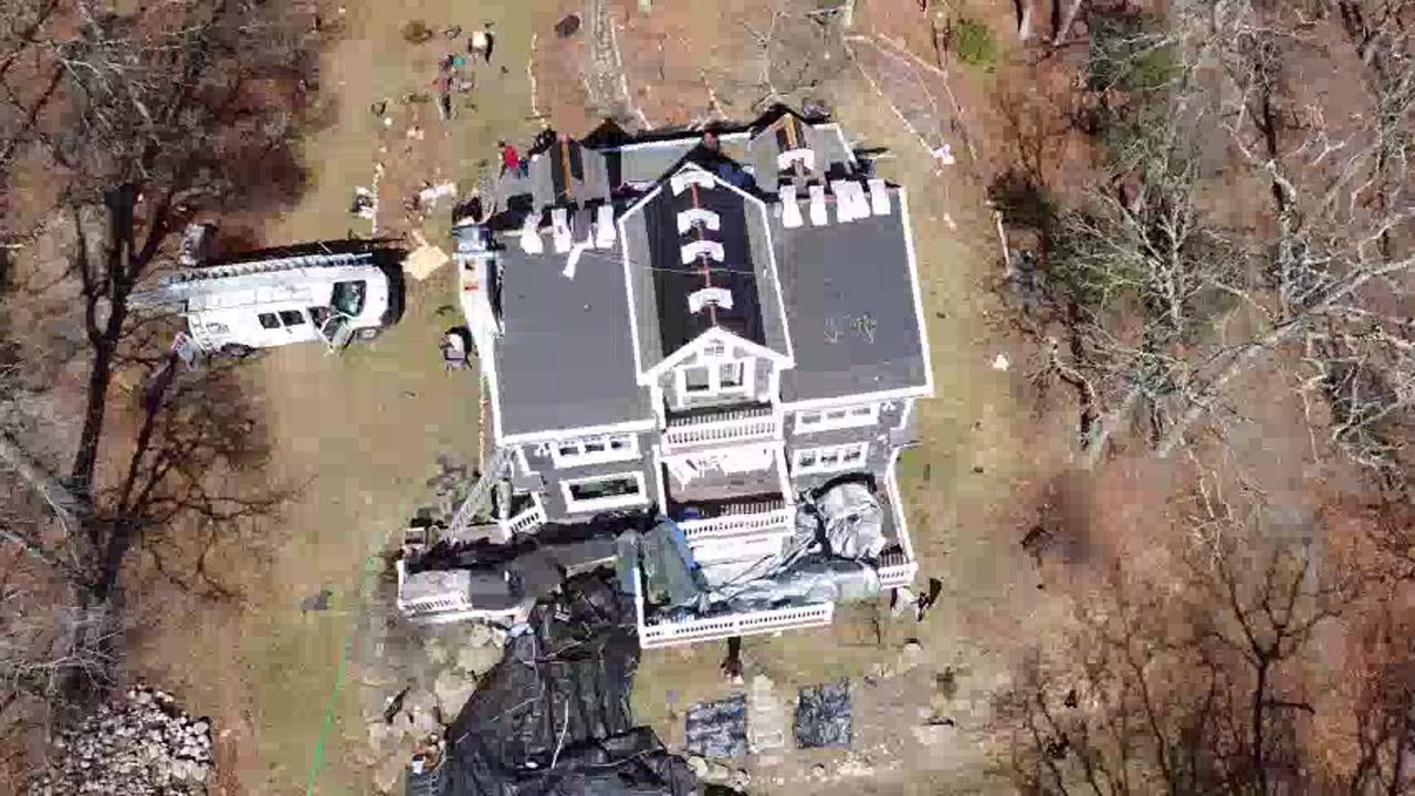 Aerial view of a large gray mansion with a driveway, parked vehicle, and leafless trees around it