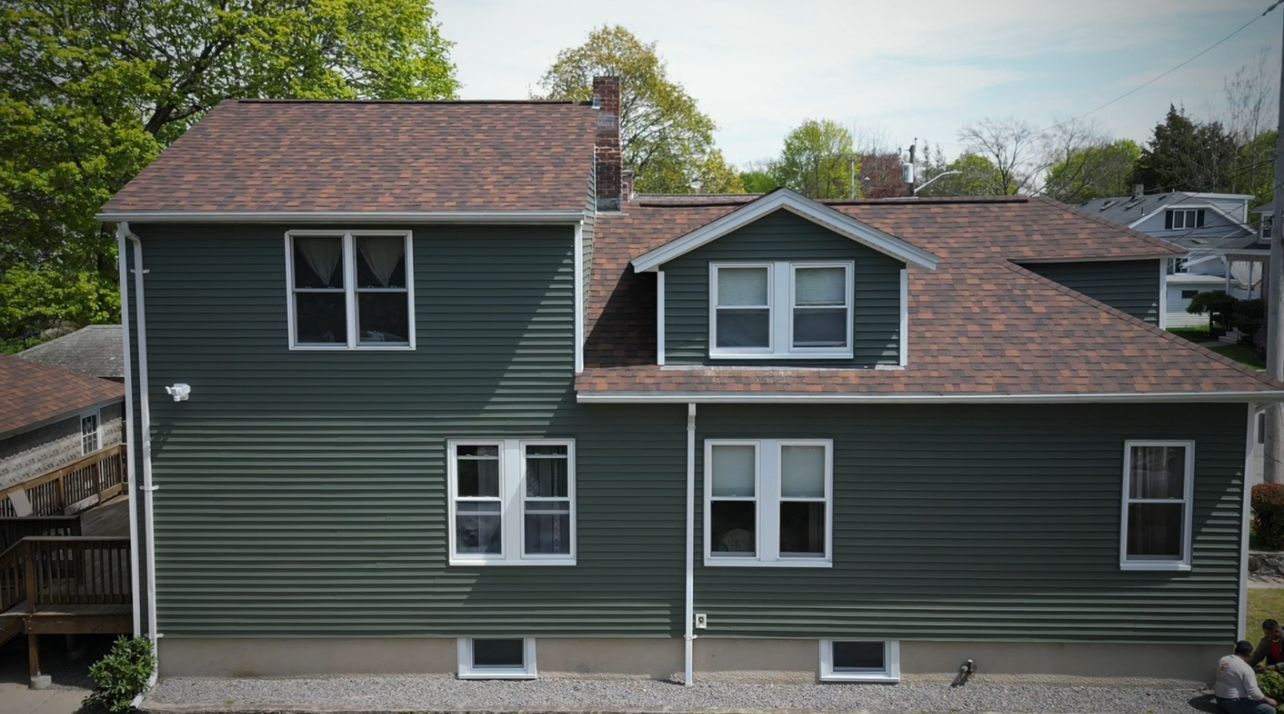 Dark green two-story house with multiple windows, brown shingle roof, and trees in the background