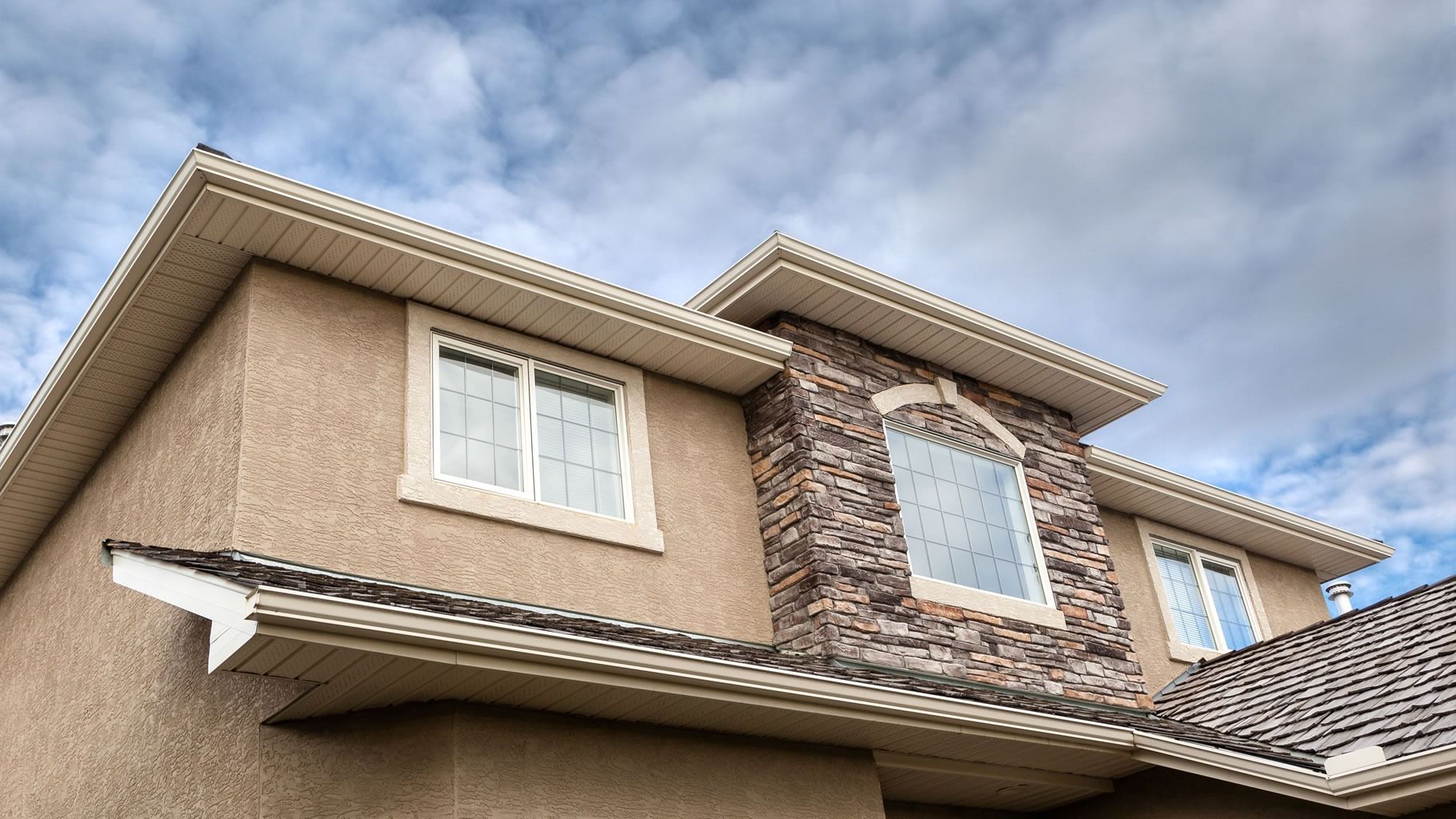 Two-story beige house exterior with white-trimmed windows and a tiled roof under a cloudy sky