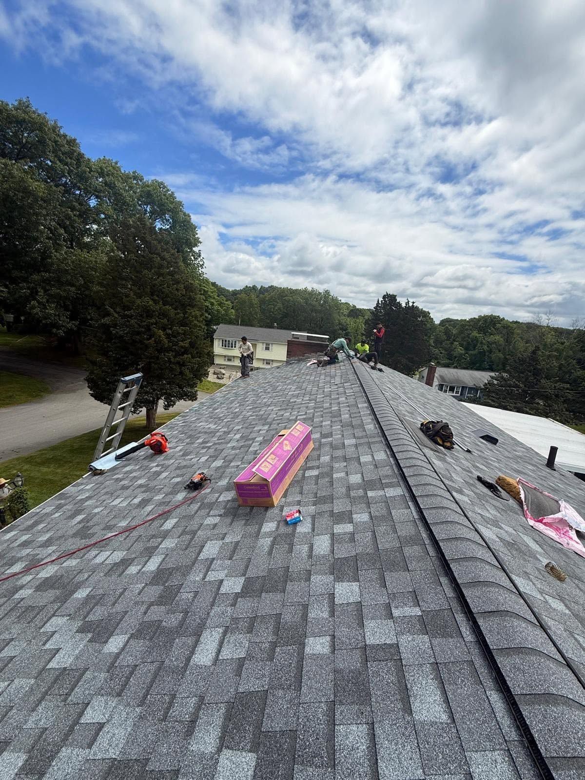 Roofing workers install gray shingles on a steep house roof under a cloudy sky.