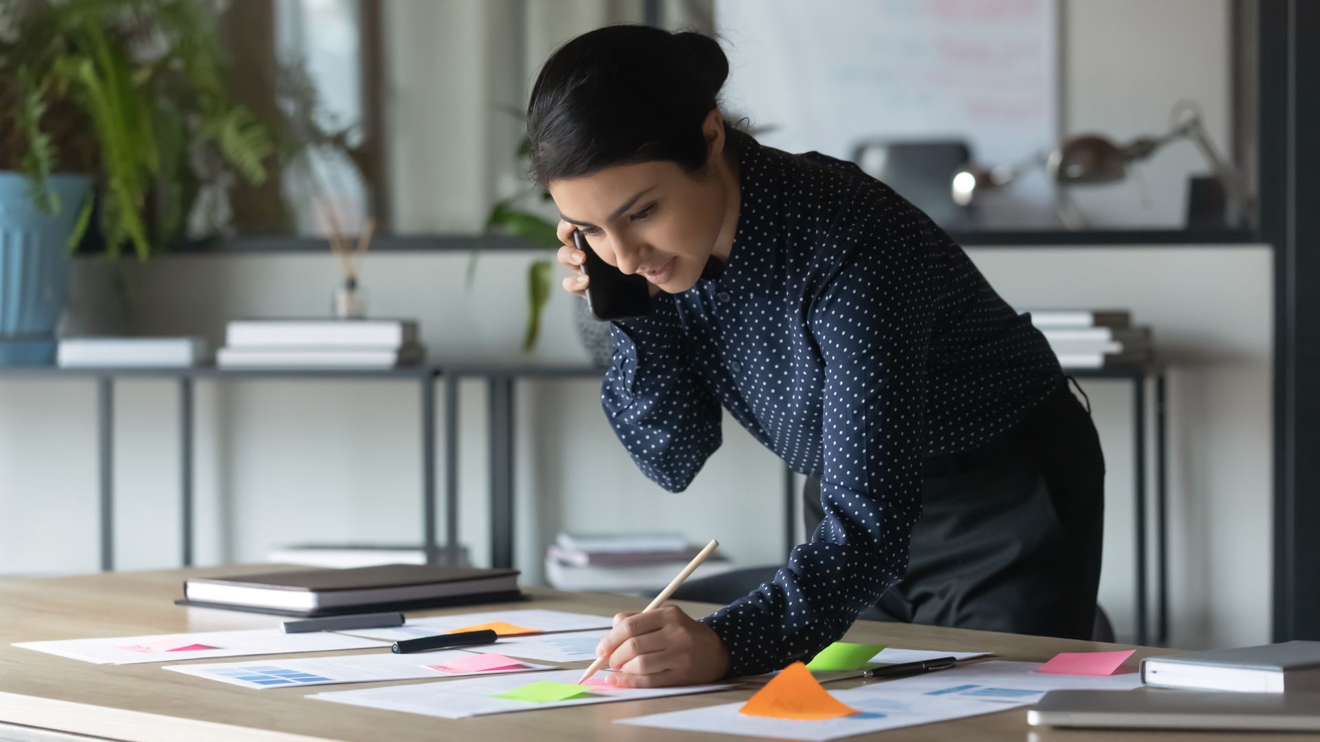 Person leaning over a desk, reviewing papers and writing in a bright office.