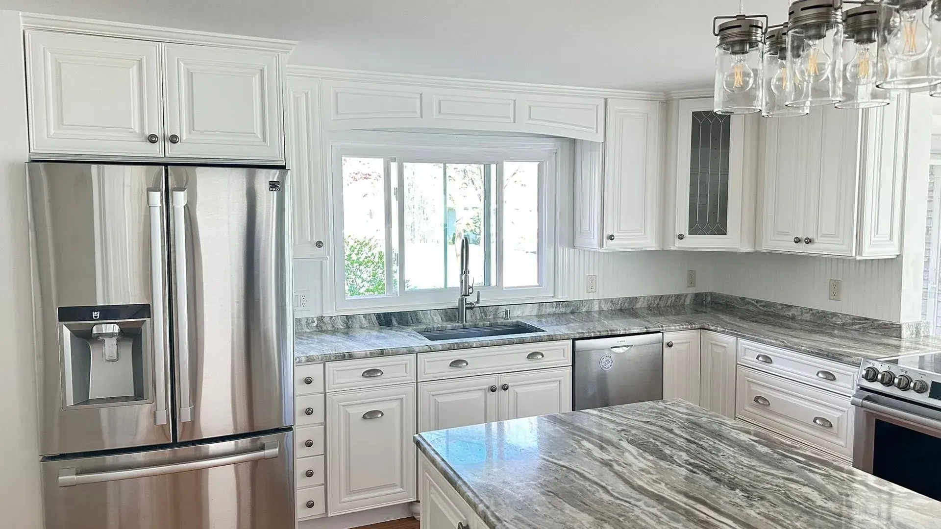 Bright white kitchen with stainless steel appliances, granite island, and large window over the sink