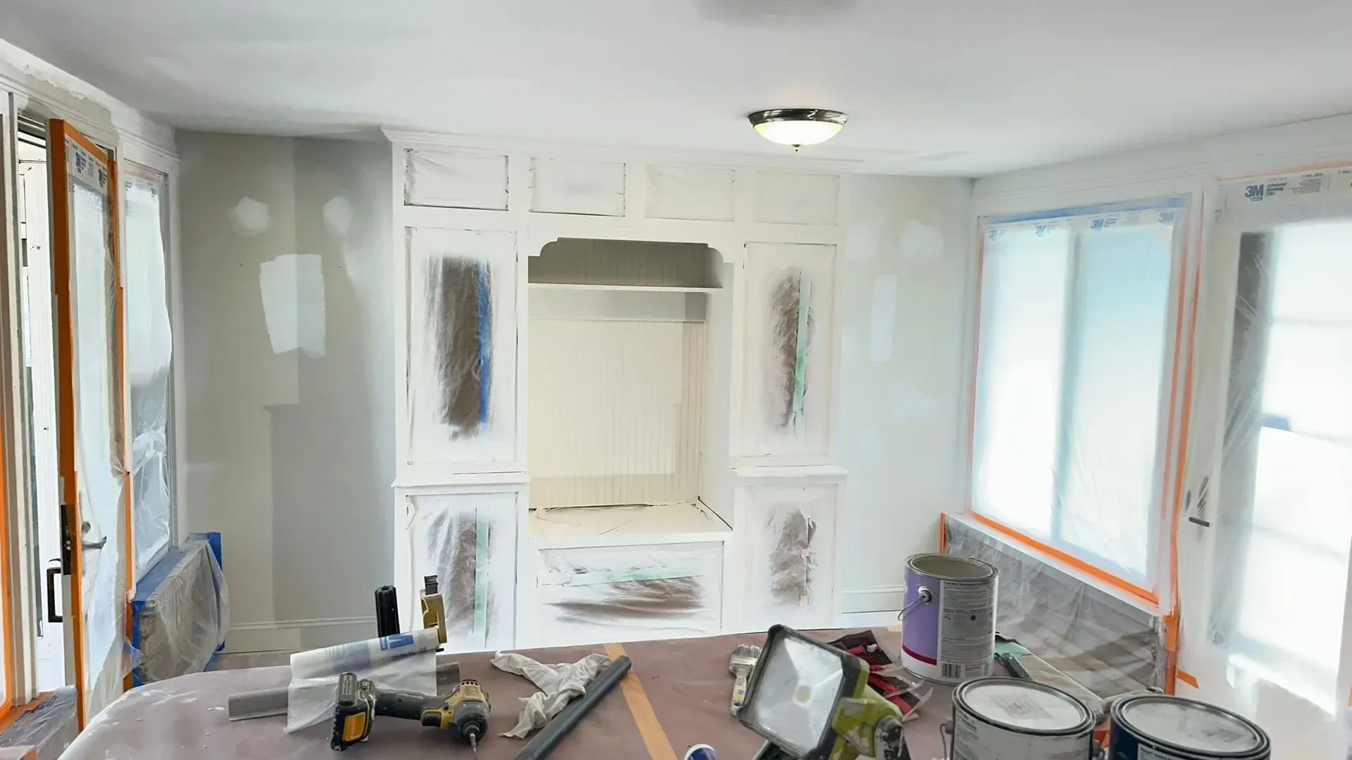 Bright, unfinished living room with white built-in cabinets, a tarp-covered table, and painting supplies.