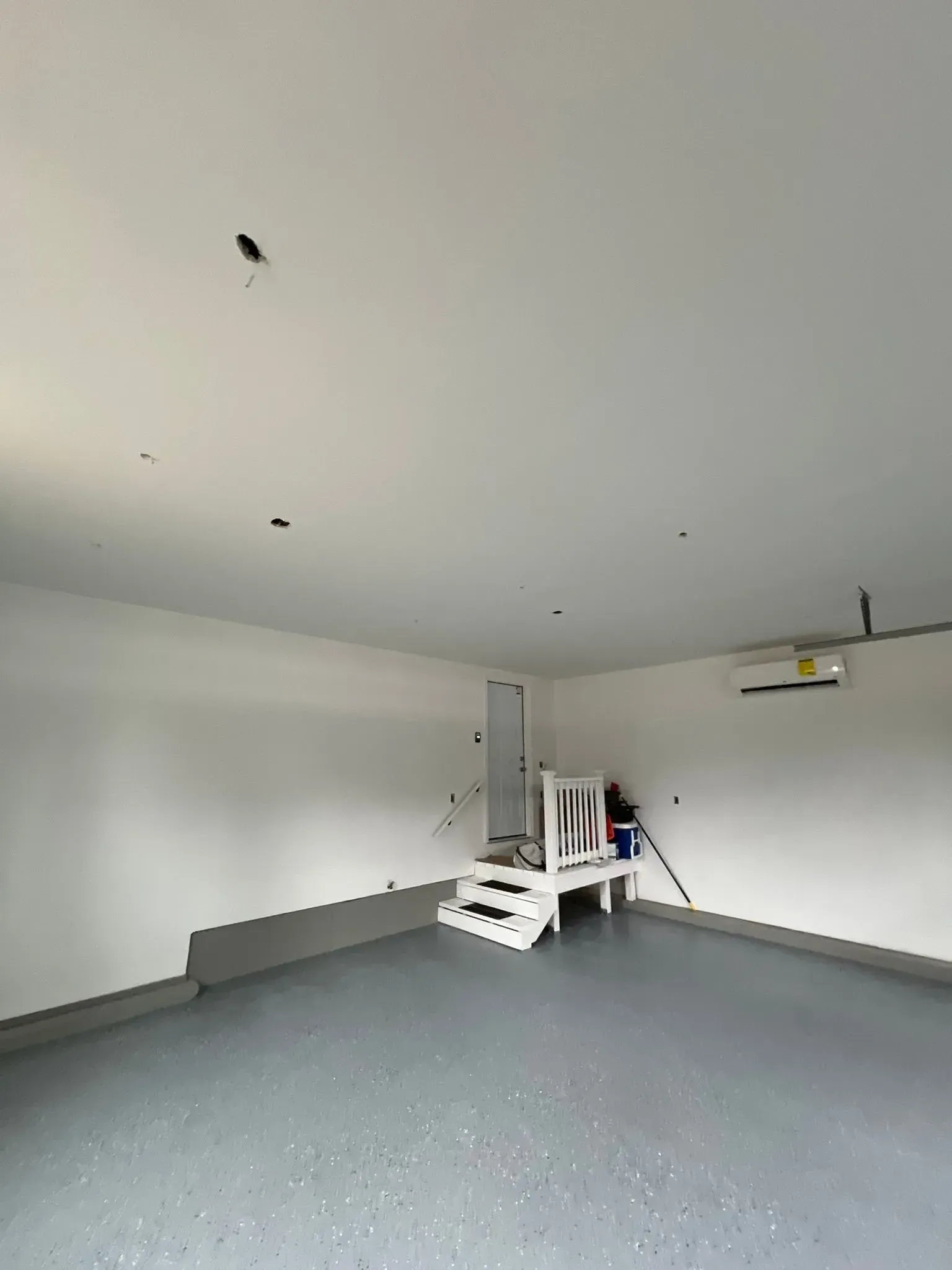 Empty garage under renovation with white walls, gray floor, and a ladder near the back wall