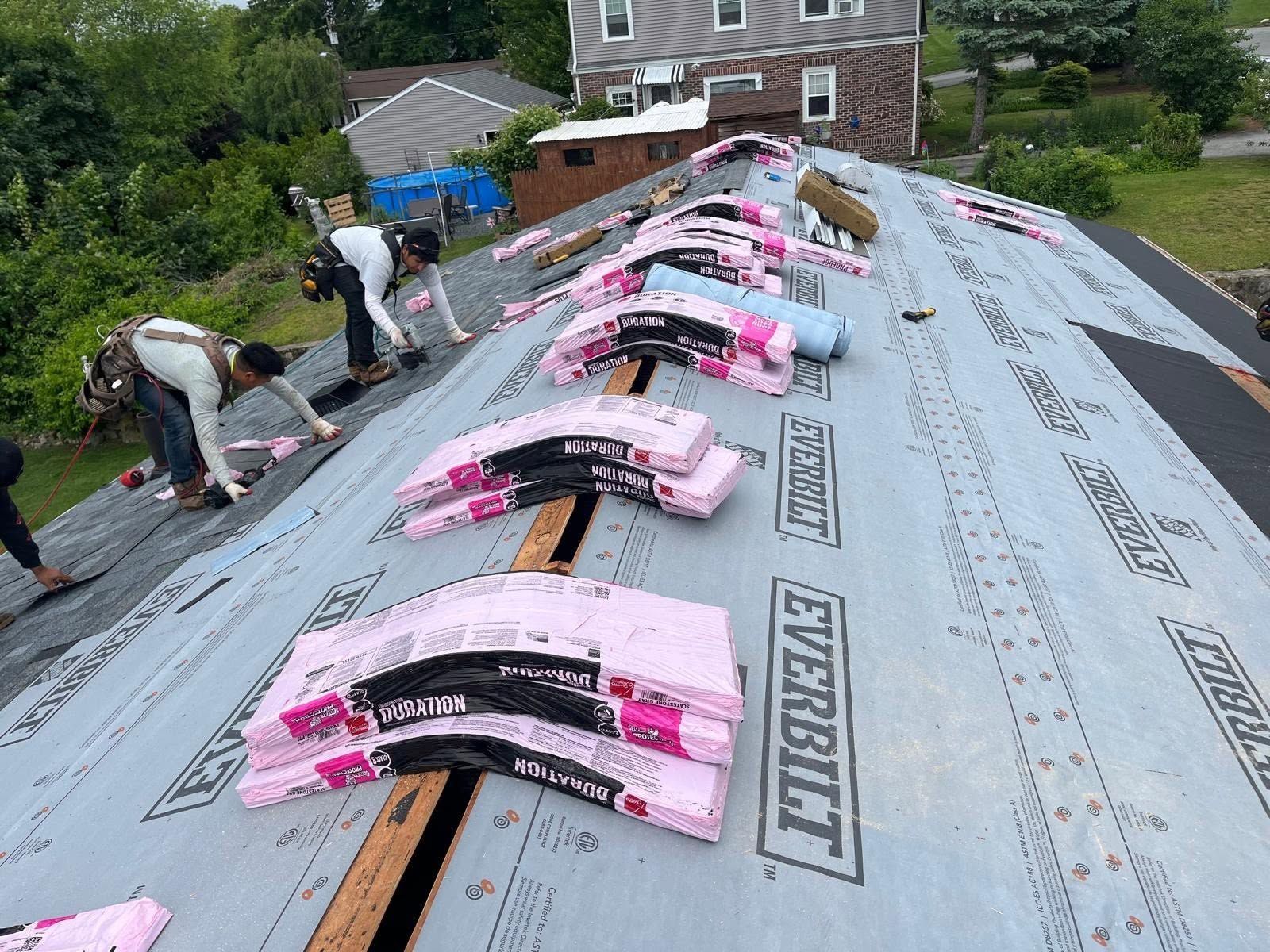 Workers installing pink roofing shingles on a house roof with gray underlayment and trees in the background