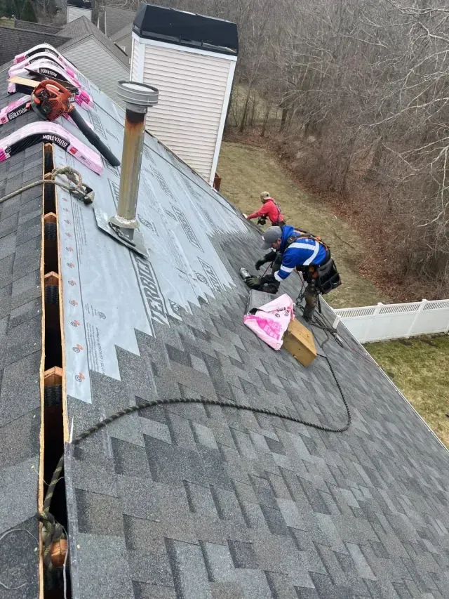 Roofers installing gray shingles on a steep roof with a chimney, using a pink tarp near the ridge.