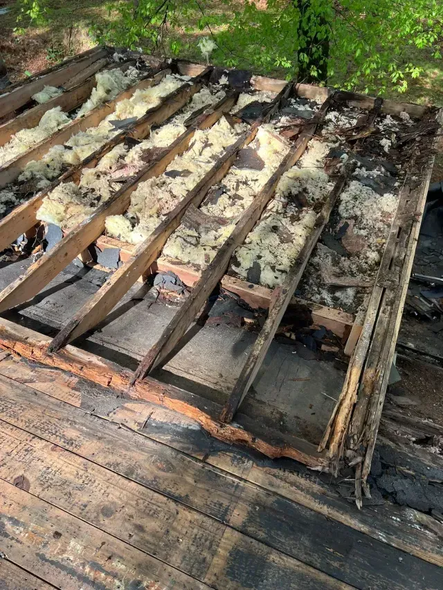 Damaged wooden deck with warped beams and missing boards, surrounded by debris and greenery