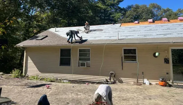 Workers installing a gray roof on a tan house with white trim and trees in the background