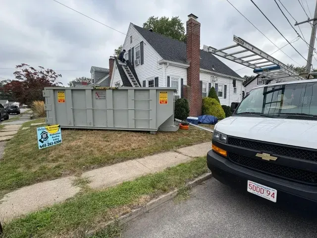 Residential driveway with a large gray dumpster beside a white house and a black van in the foreground