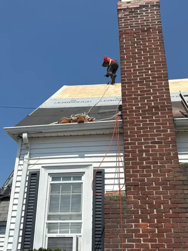 Worker on a roof near a brick chimney, repairing shingles on a white house under a clear blue sky