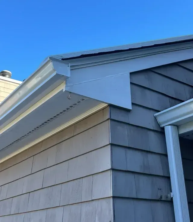 Close-up of a house roofline with white trim and gray shingle siding against a blue sky