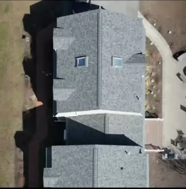 Aerial view of a gray-shingled house roof with skylights and a driveway beside it.
