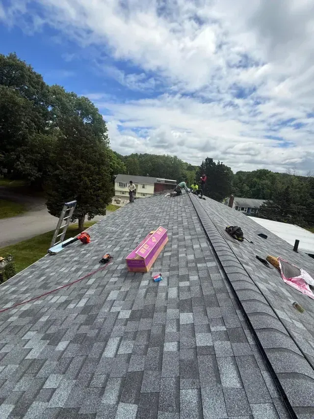 Roof under construction with gray shingles, pink materials, and workers on a partly finished roof under cloudy sky